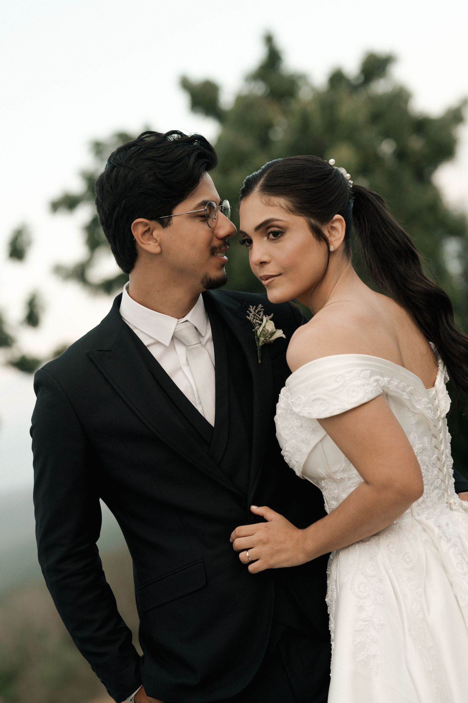 A bride in an off-shoulder white gown and groom in a black suit share an intimate moment outdoors.