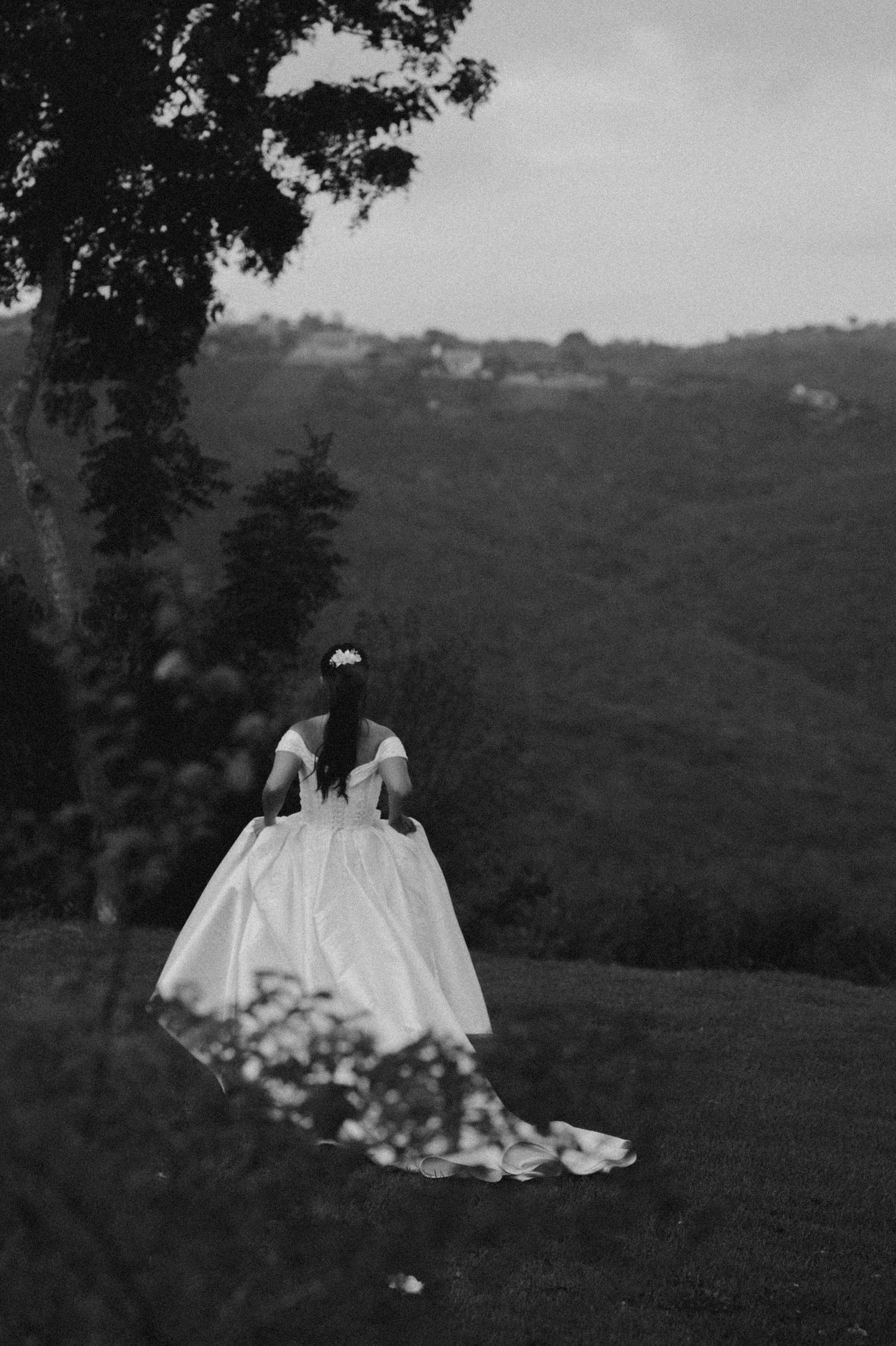 Black and white photo of a bride in a flowing white gown standing on a hillside overlooking lush green mountains.