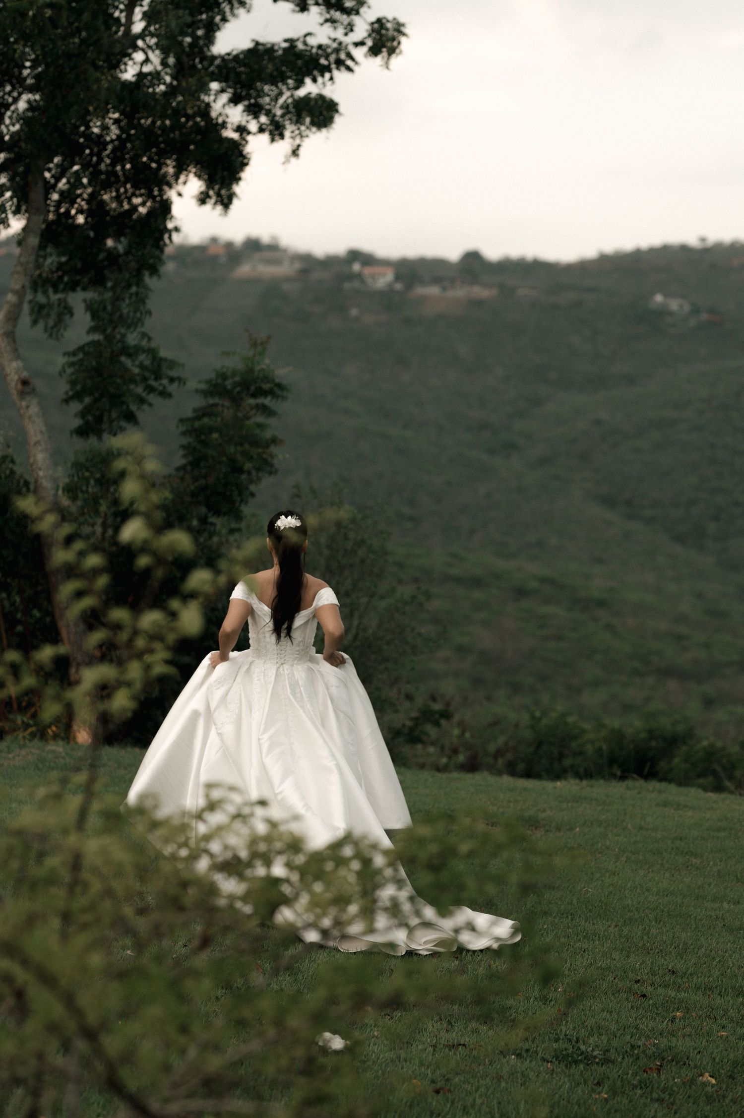 A bride in an elegant white ball gown with a train stands on a green hillside with scenic mountain views.