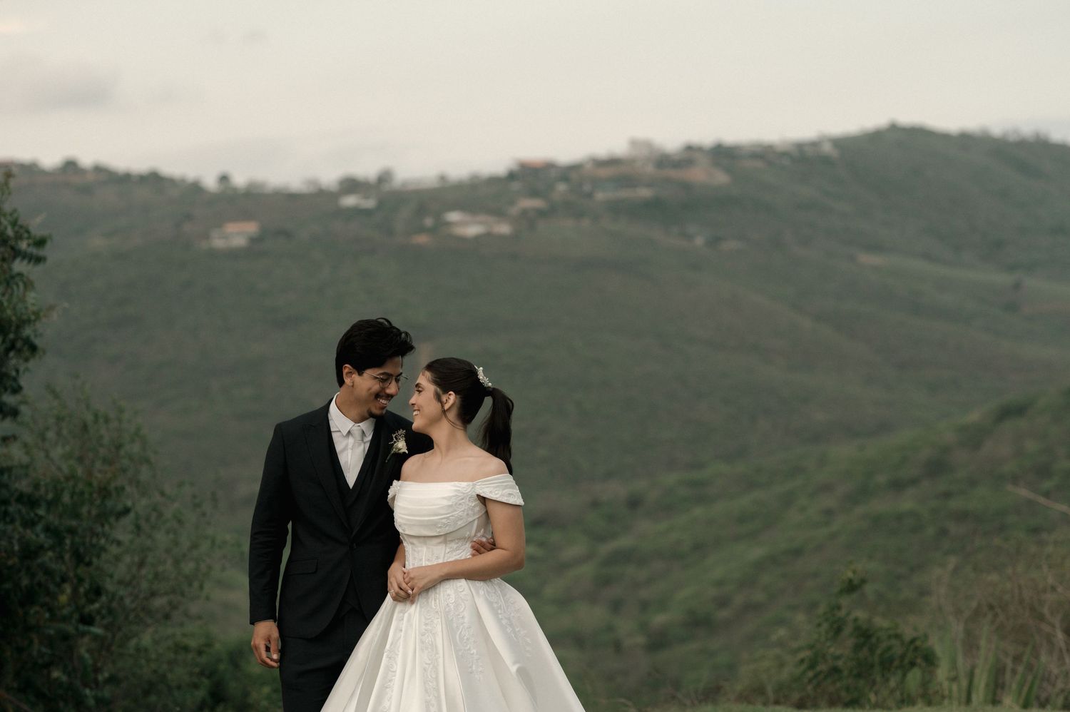 A couple in wedding attire shares a romantic moment with lush green hills in the background.