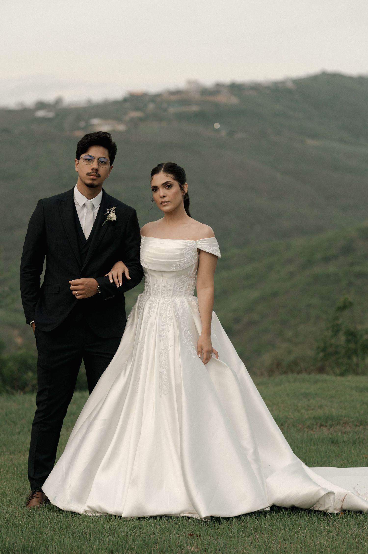 Elegant bride in off-shoulder white ball gown and groom in black suit pose outdoors with green hills backdrop.