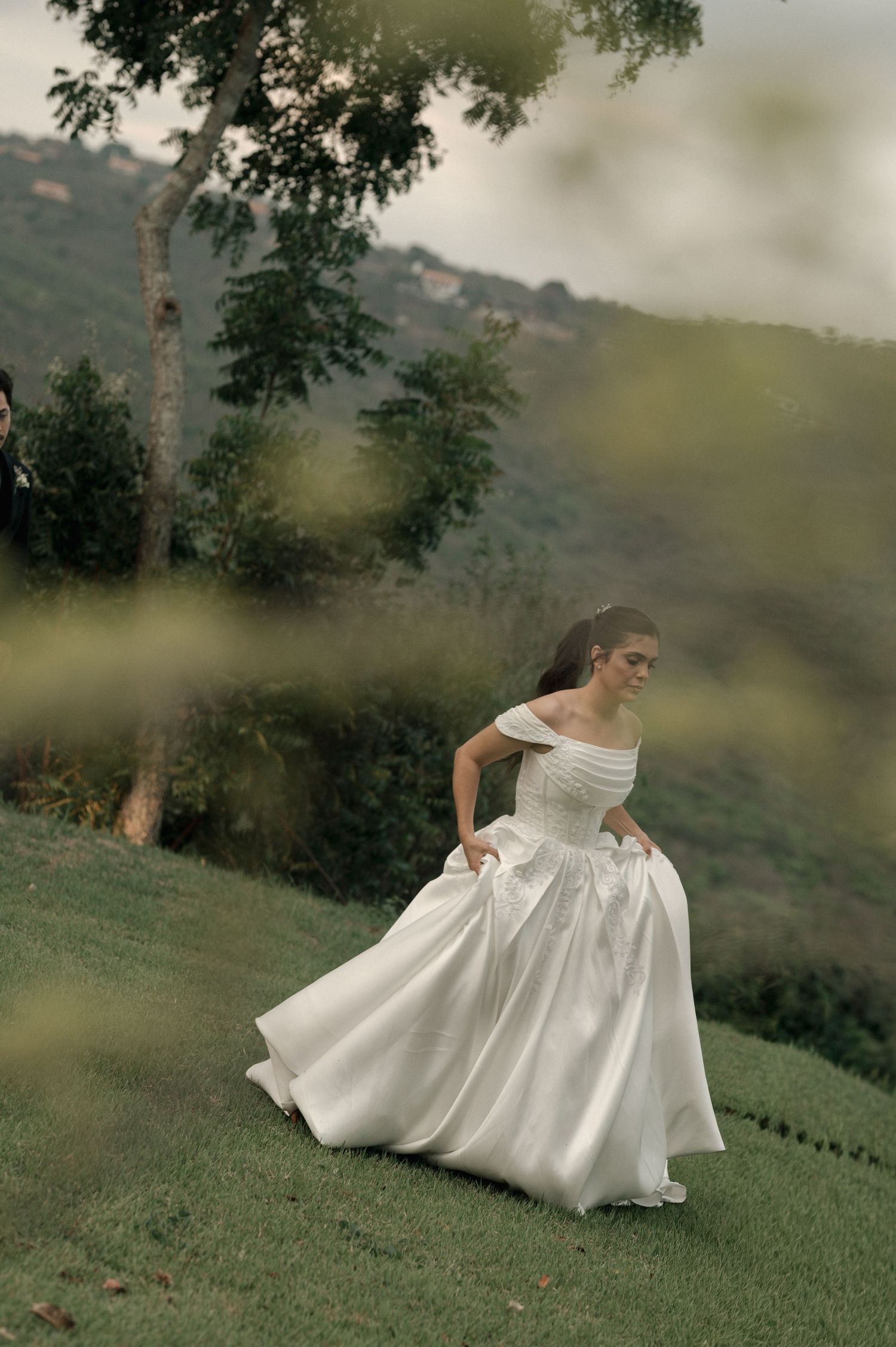 Bride in off-shoulder white ball gown walking on lush green hillside with trees and mountains in background.