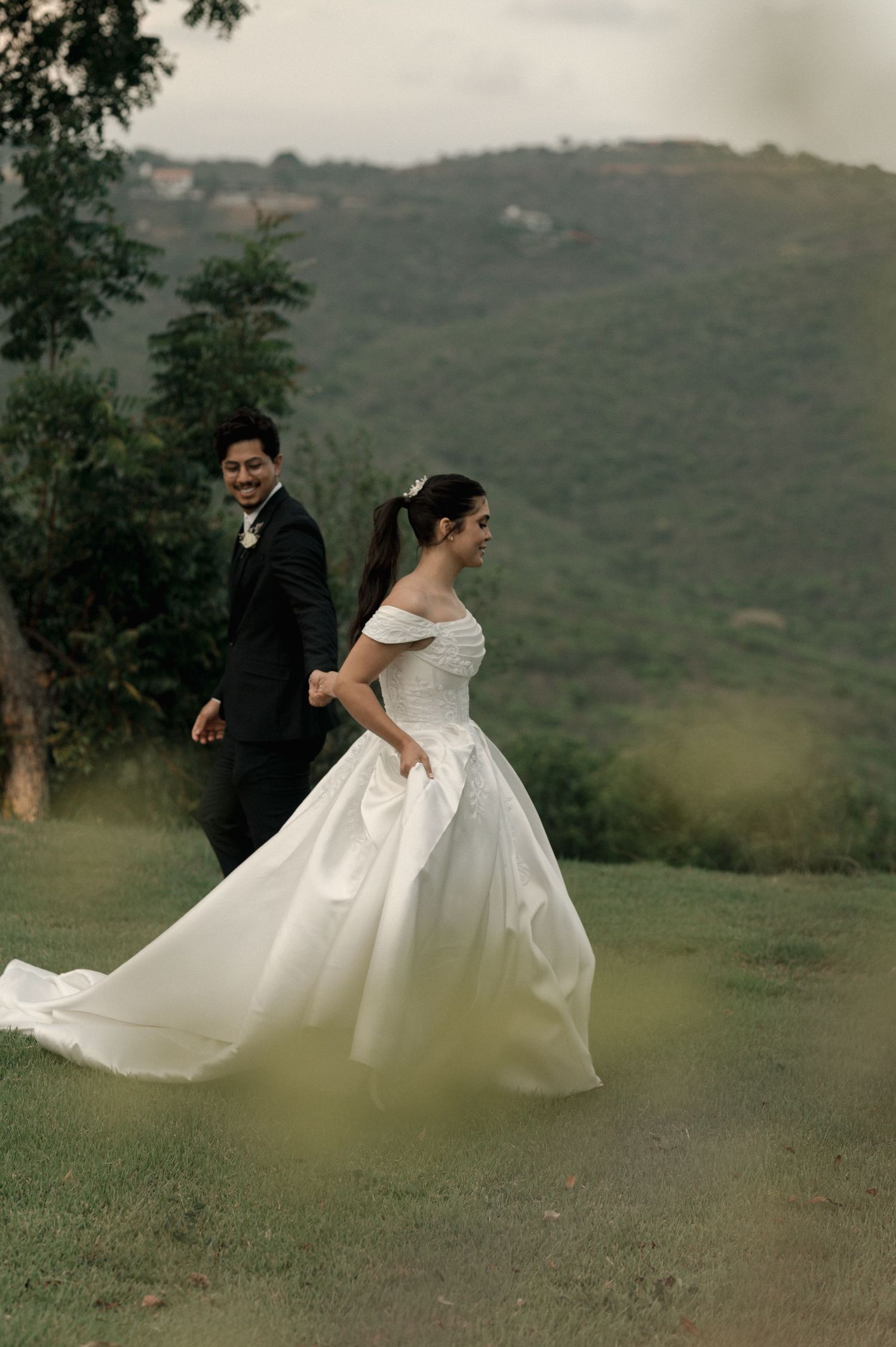 Bride in off-shoulder ball gown and groom in black suit walk through green hillside landscape during outdoor wedding photoshoot.