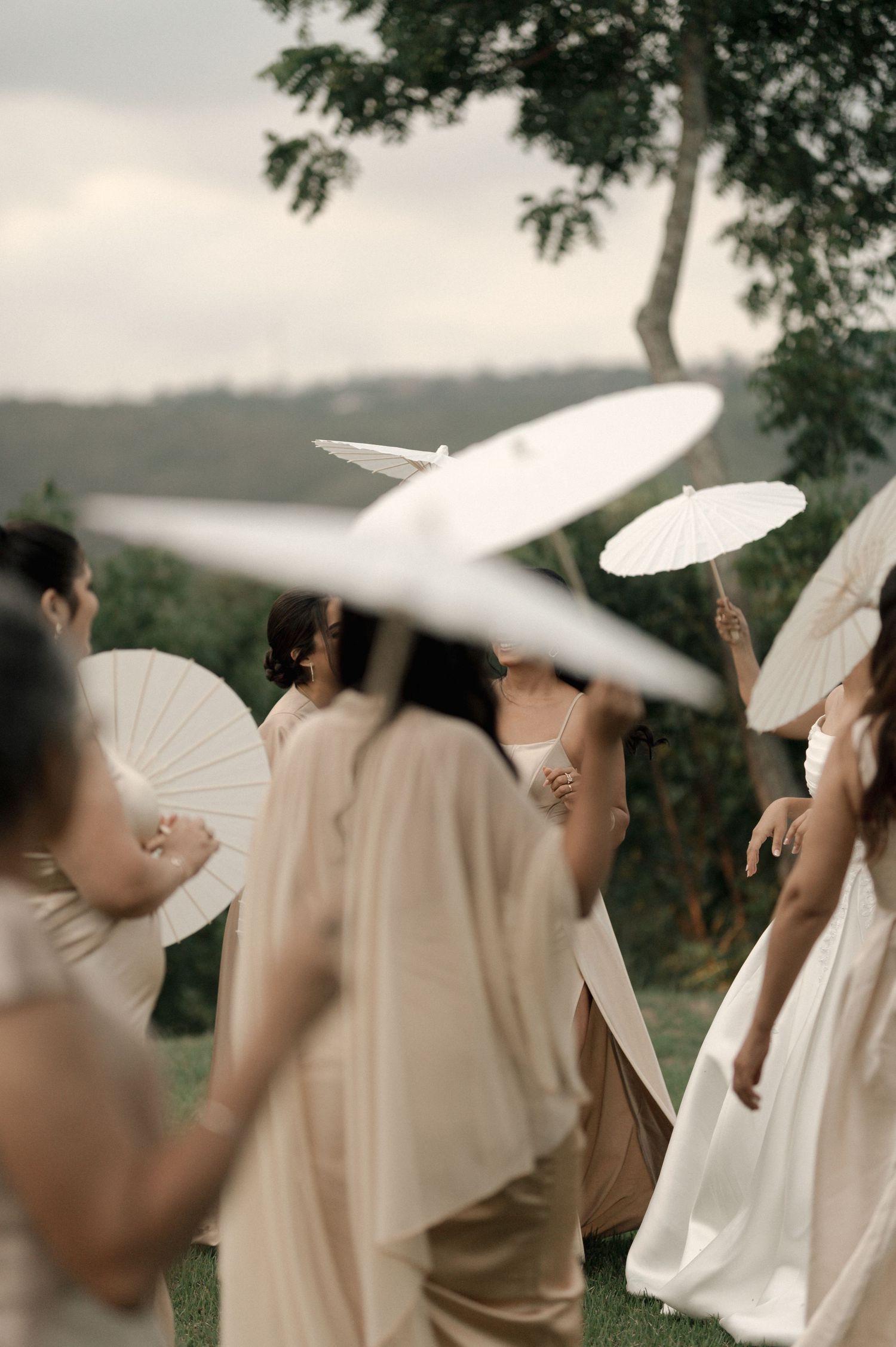 Bridesmaids in champagne gowns holding white paper parasols outdoors at an elegant garden wedding ceremony.