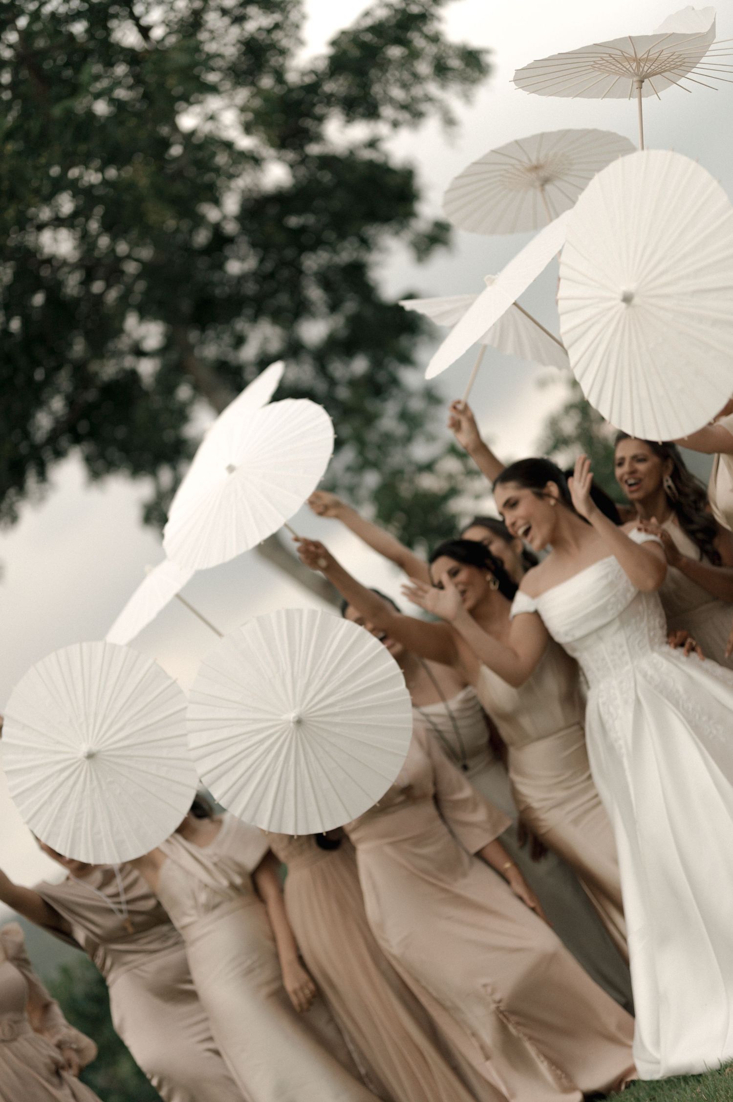 Bride and bridesmaids in white and champagne gowns joyfully twirling white paper parasols outdoors at a wedding celebration.