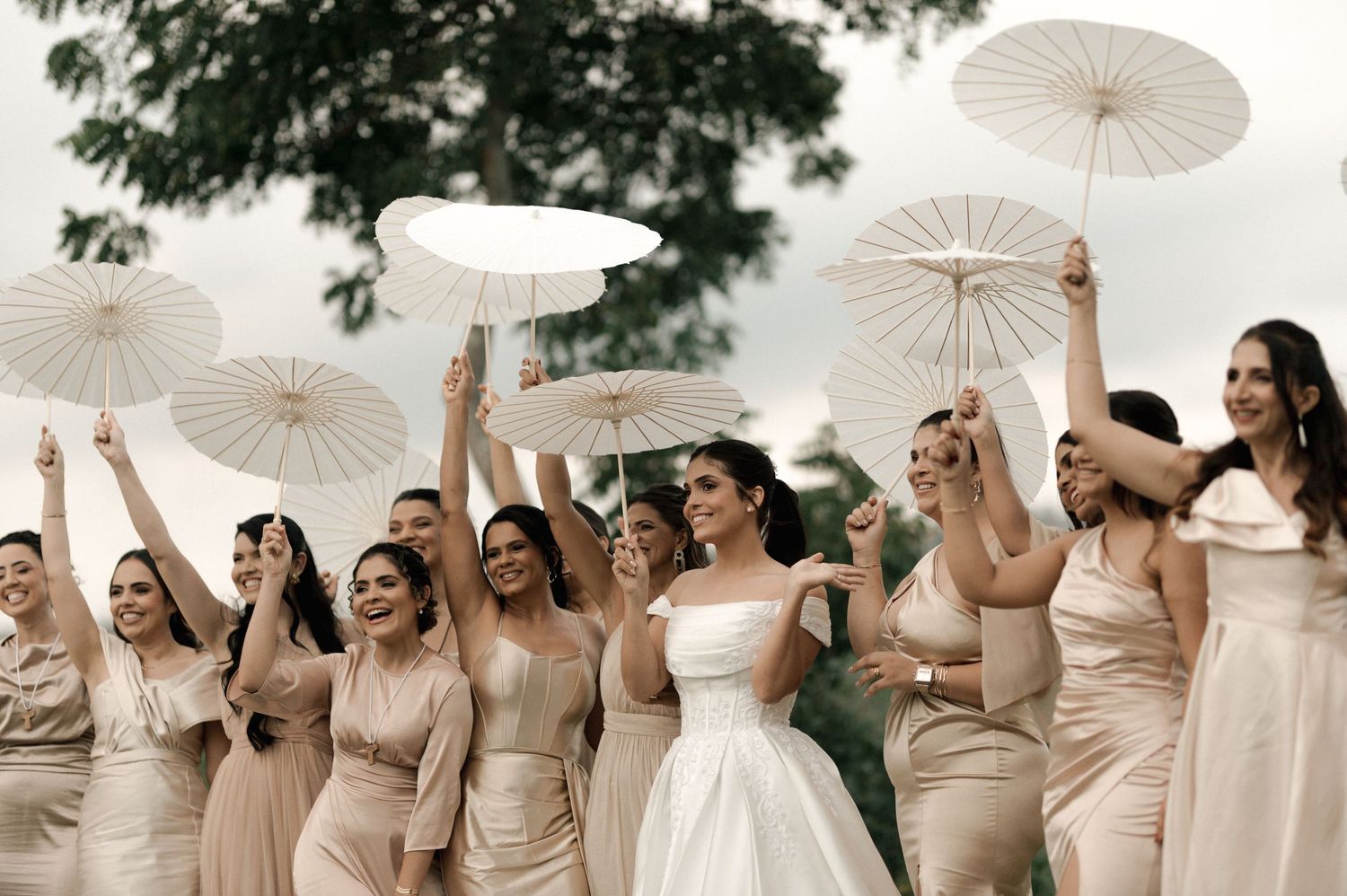Bride and bridesmaids in champagne dresses celebrate outdoors, joyfully raising white paper parasols.