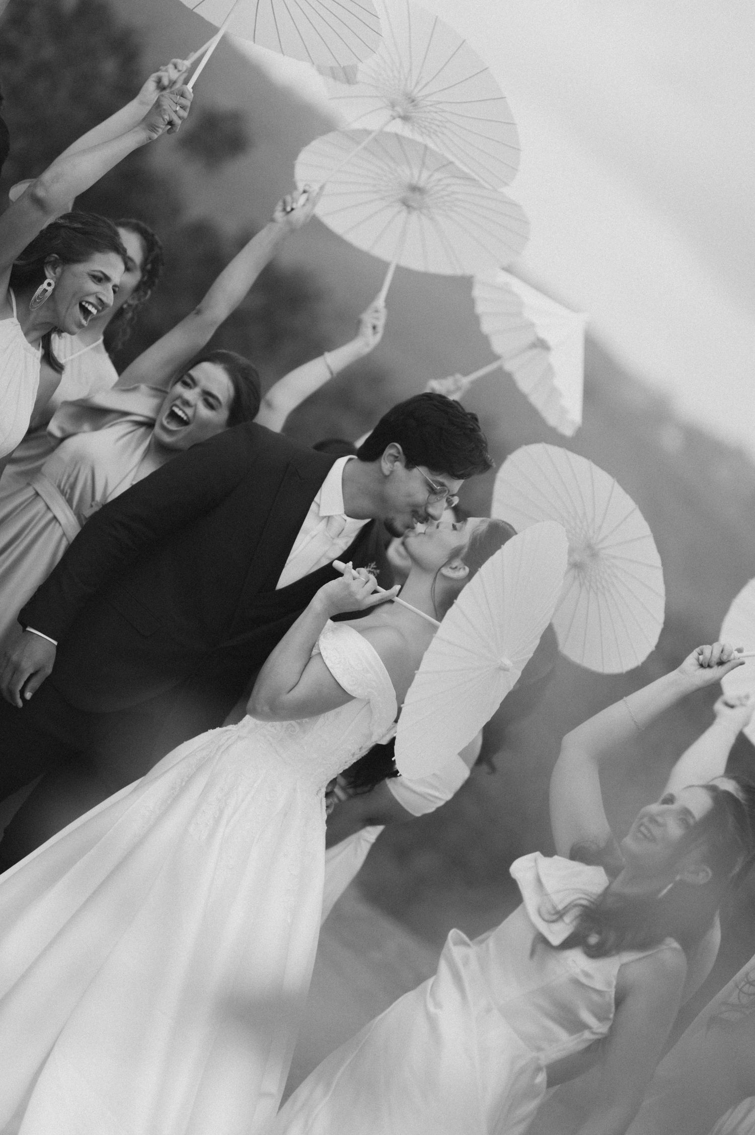 Black and white wedding photo of couple kissing surrounded by joyful bridesmaids holding white parasols.
