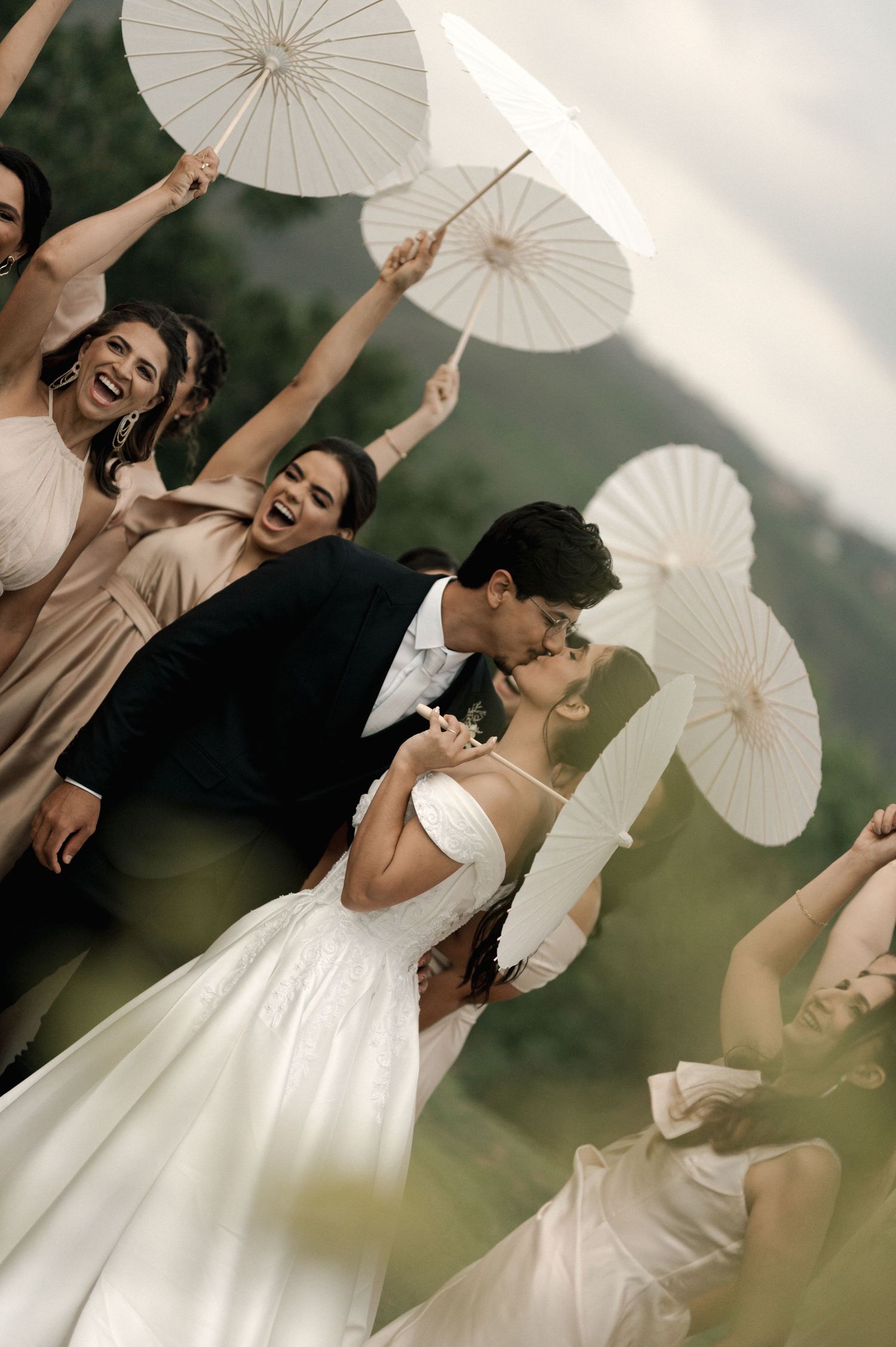 Bride and groom share a kiss surrounded by joyful bridesmaids holding white parasols outdoors.