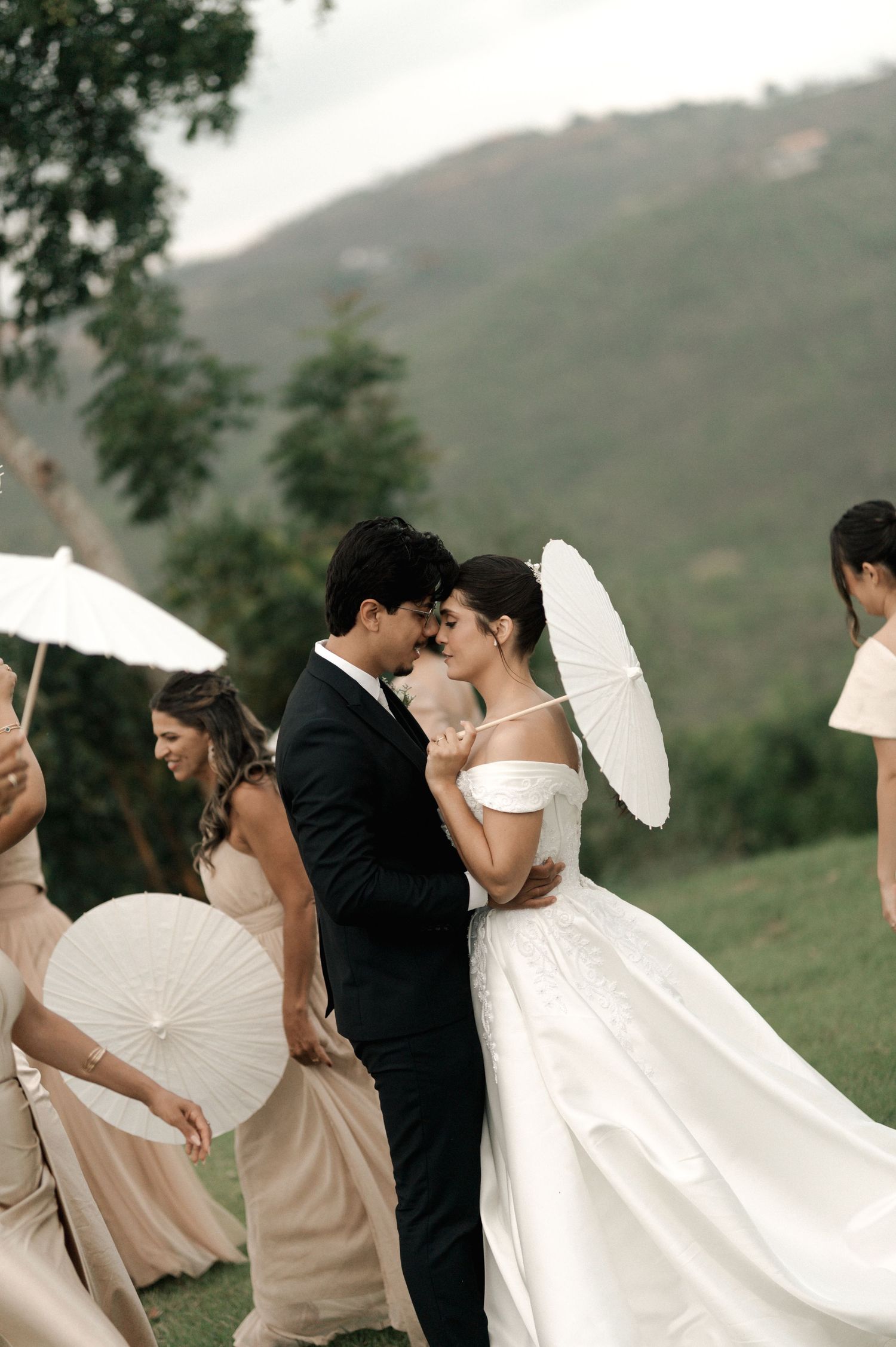 Bride and groom share romantic moment at outdoor wedding ceremony with bridesmaids holding white parasols and mountain backdrop.