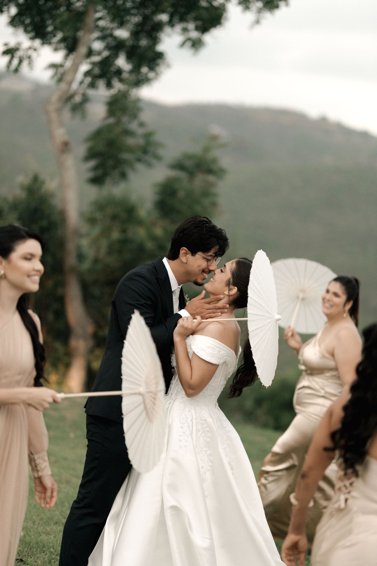 Bride and groom share a kiss outdoors surrounded by bridesmaids holding white parasols with mountain views.
