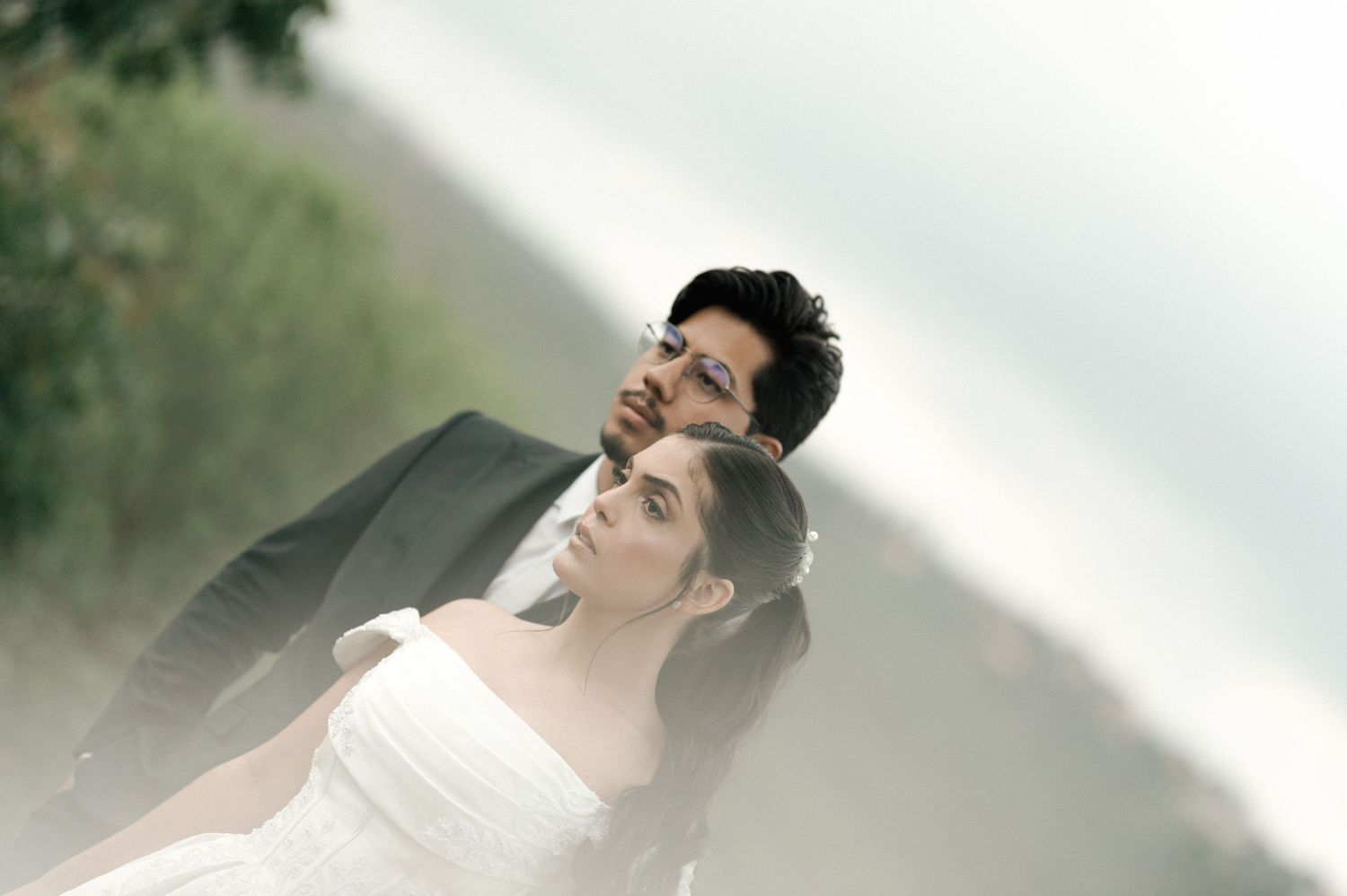 Romantic wedding couple posing outdoors, groom in dark suit behind bride in white strapless dress, misty scenic background.