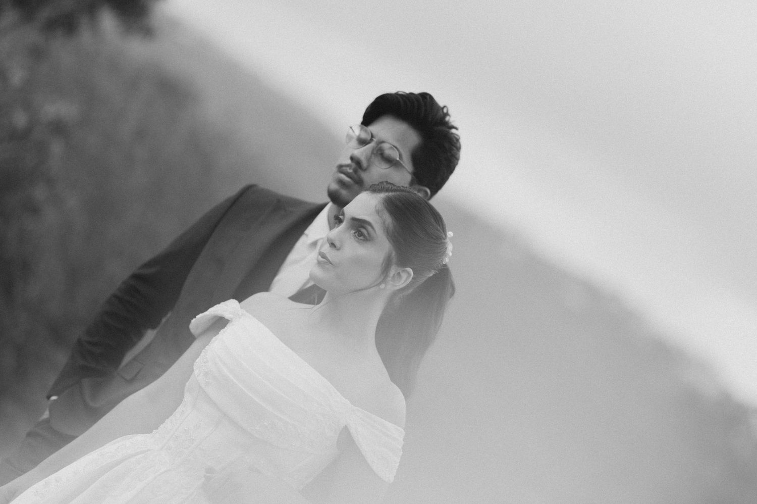 Black and white wedding photo of elegant couple, bride in off-shoulder gown, groom in suit, romantic outdoor portrait.