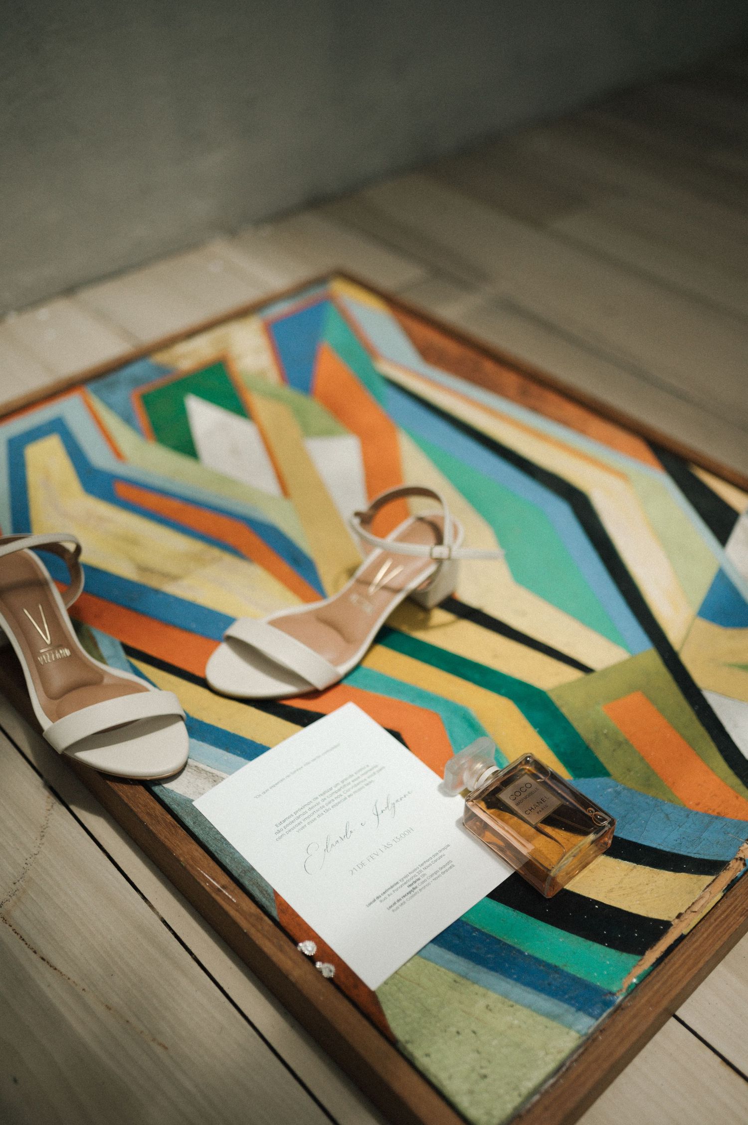 Bridal flat sandals, perfume bottle, and wedding invitation on a colorful geometric art tray on white wooden floor.