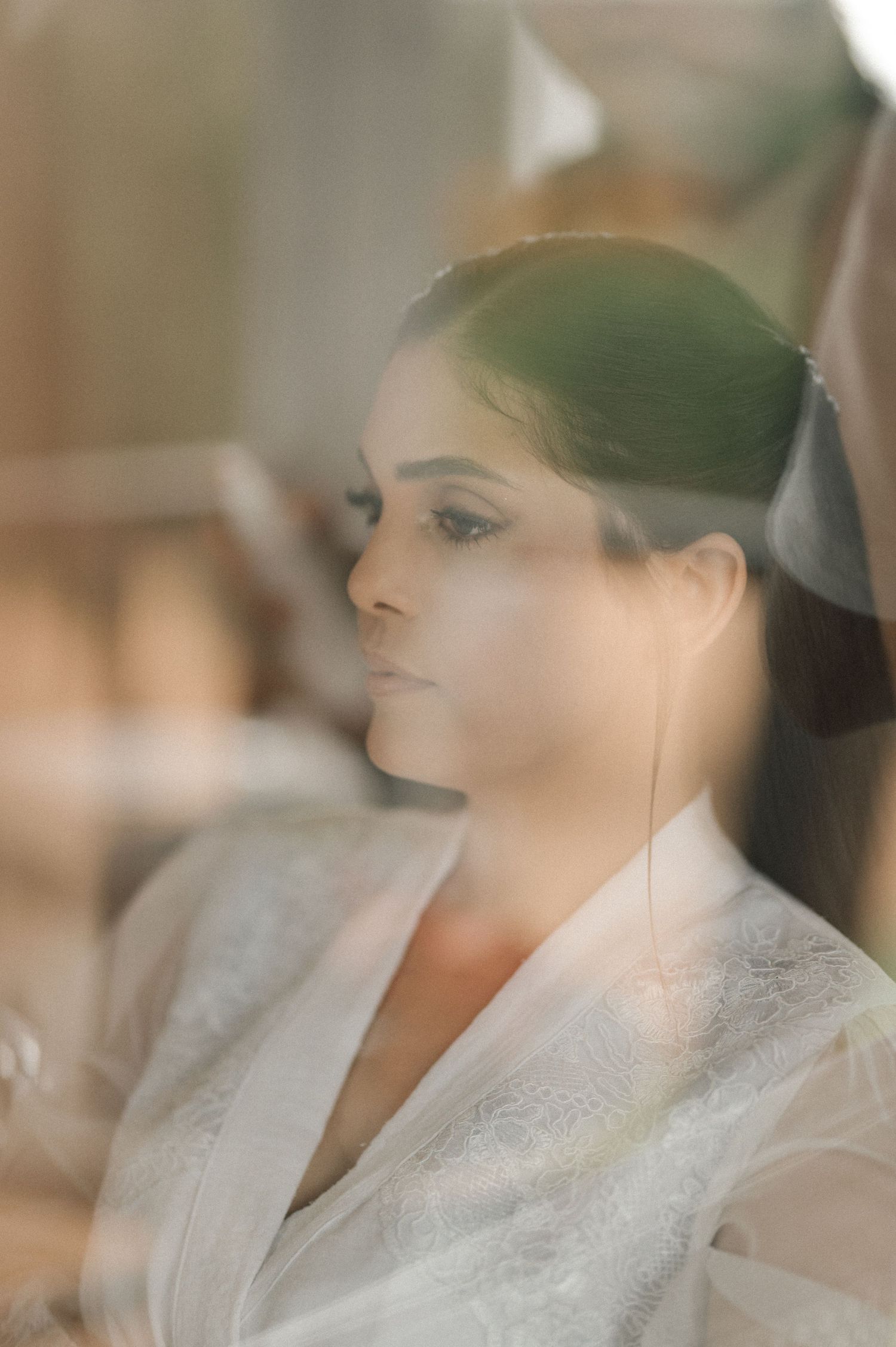 Bride in white lace robe gazing thoughtfully, captured through glass with soft reflections.