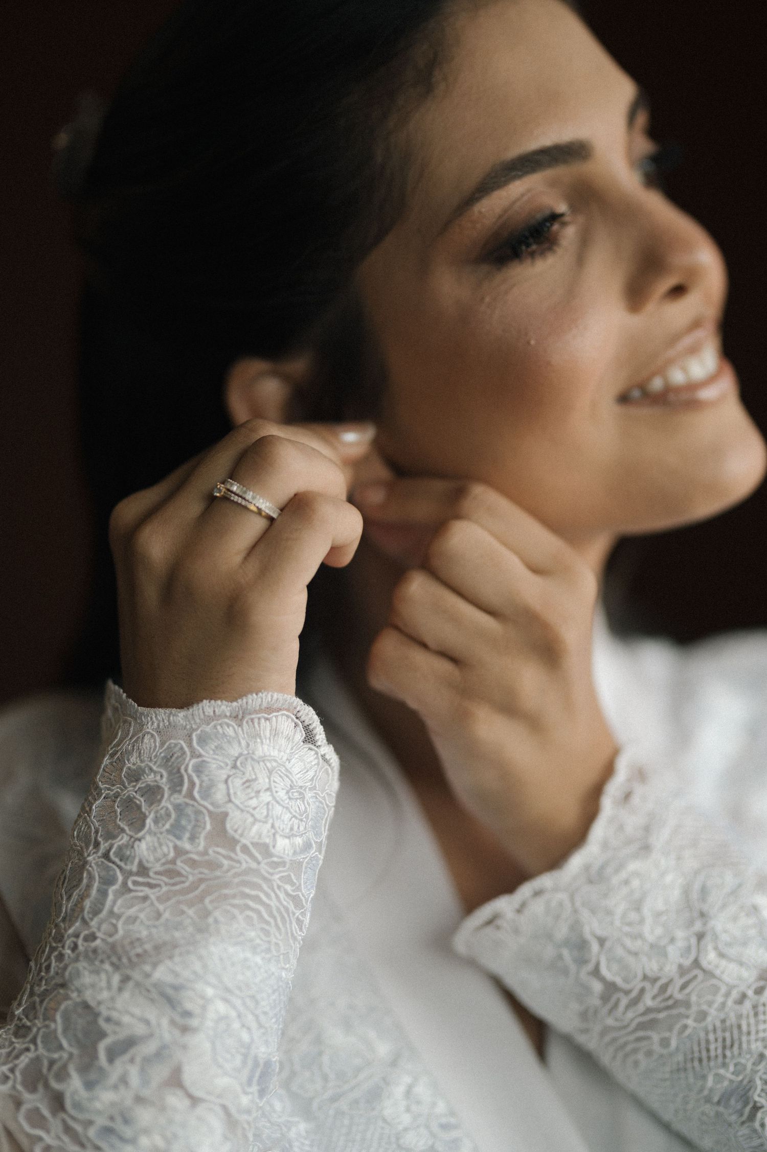 A smiling bride in lace dress putting on earrings, wearing a diamond ring, with elegant makeup on her wedding day.