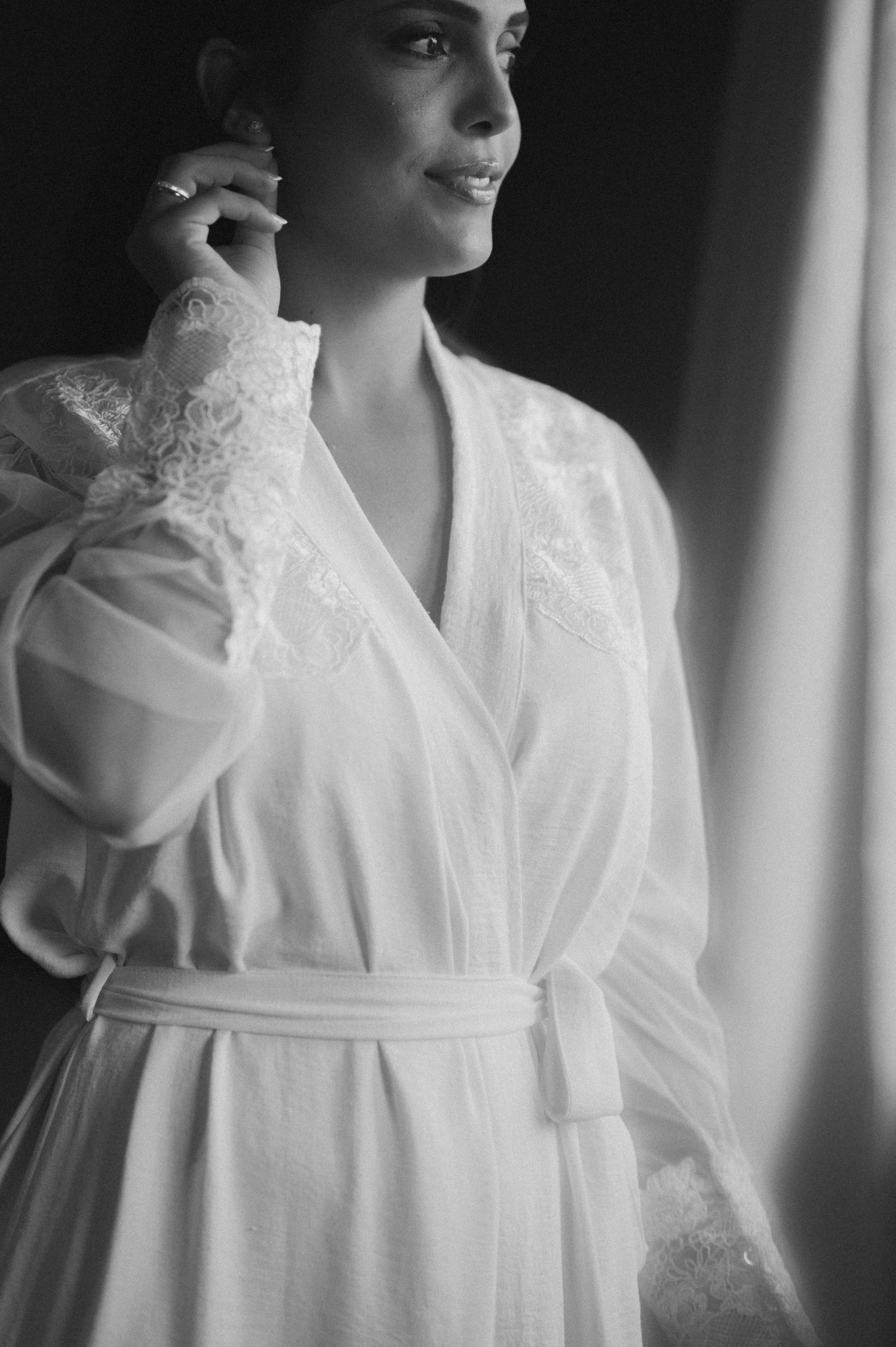 Bride in elegant white lace robe smiling softly by window in black and white photo.