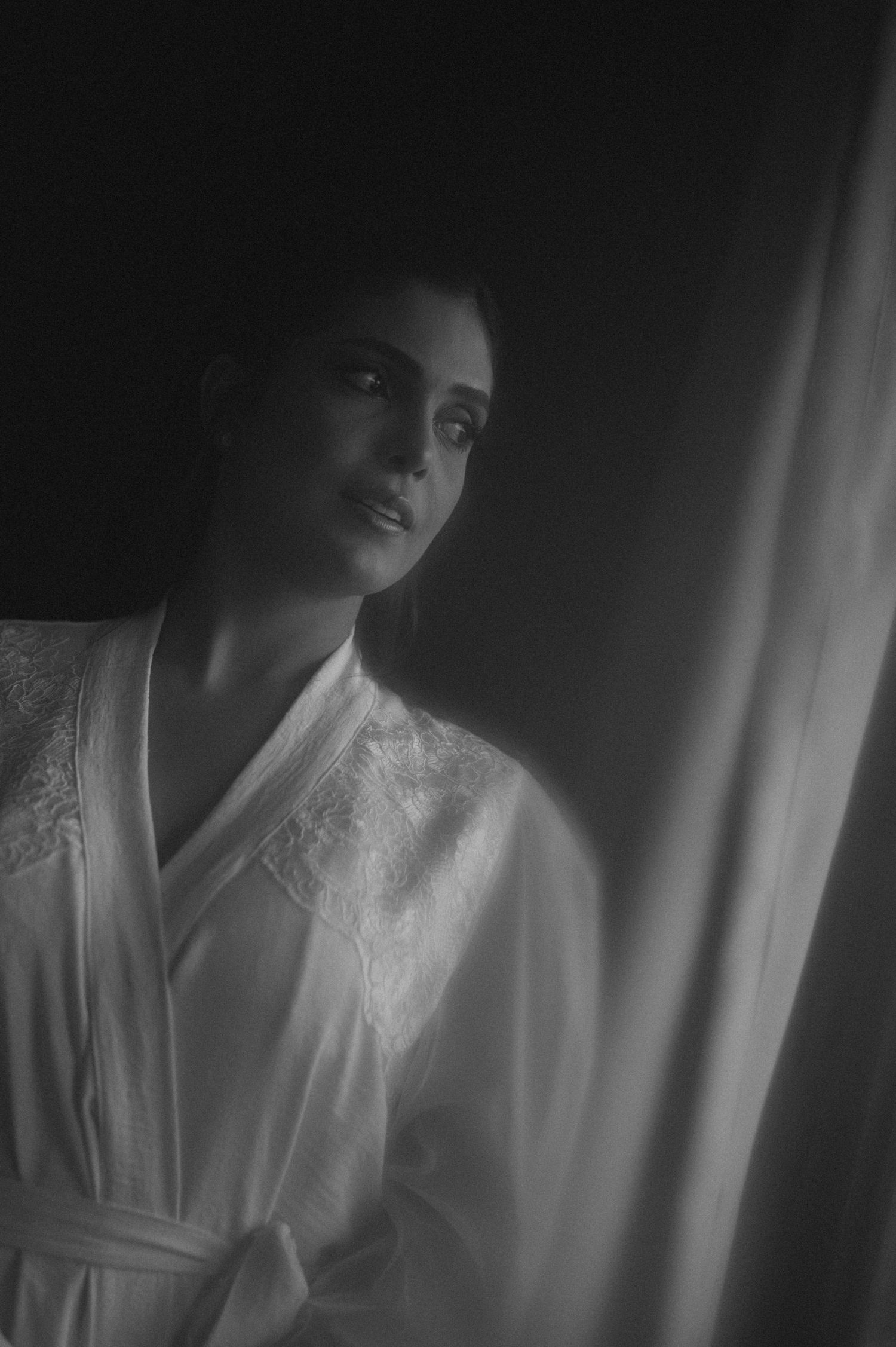 Black and white portrait of a woman in a white robe standing near a curtain in moody dramatic lighting.