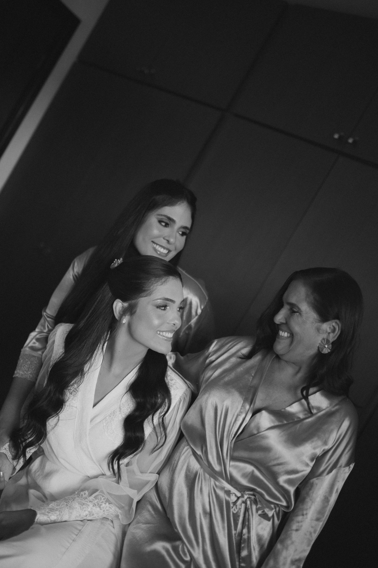 Three women in satin robes smile together in a black and white bridal getting-ready photo.