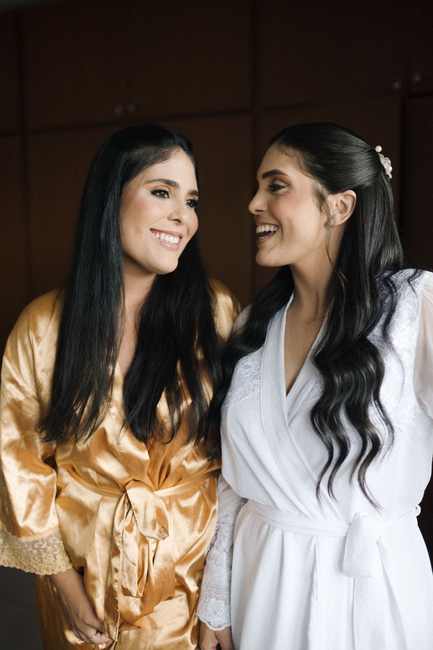 Two smiling women in satin robes, one gold and one white, share a joyful moment during bridal preparations.