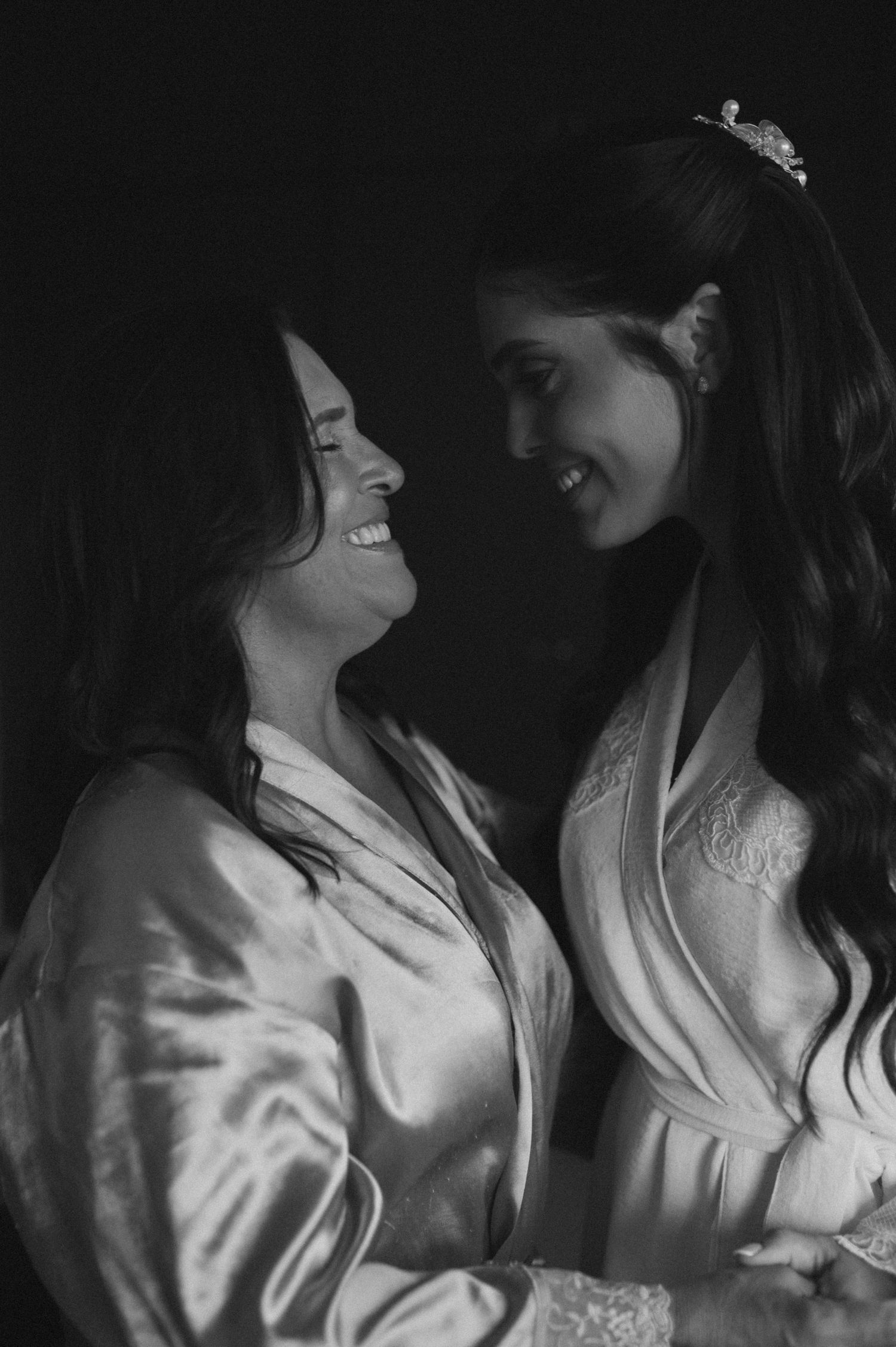Two smiling women in satin robes share a joyful moment, one wearing a tiara, in a black and white photo.