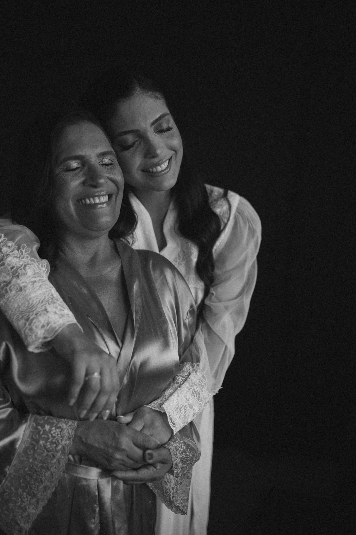 Black and white photo of two women in satin robes sharing a warm embrace, laughing joyfully together.
