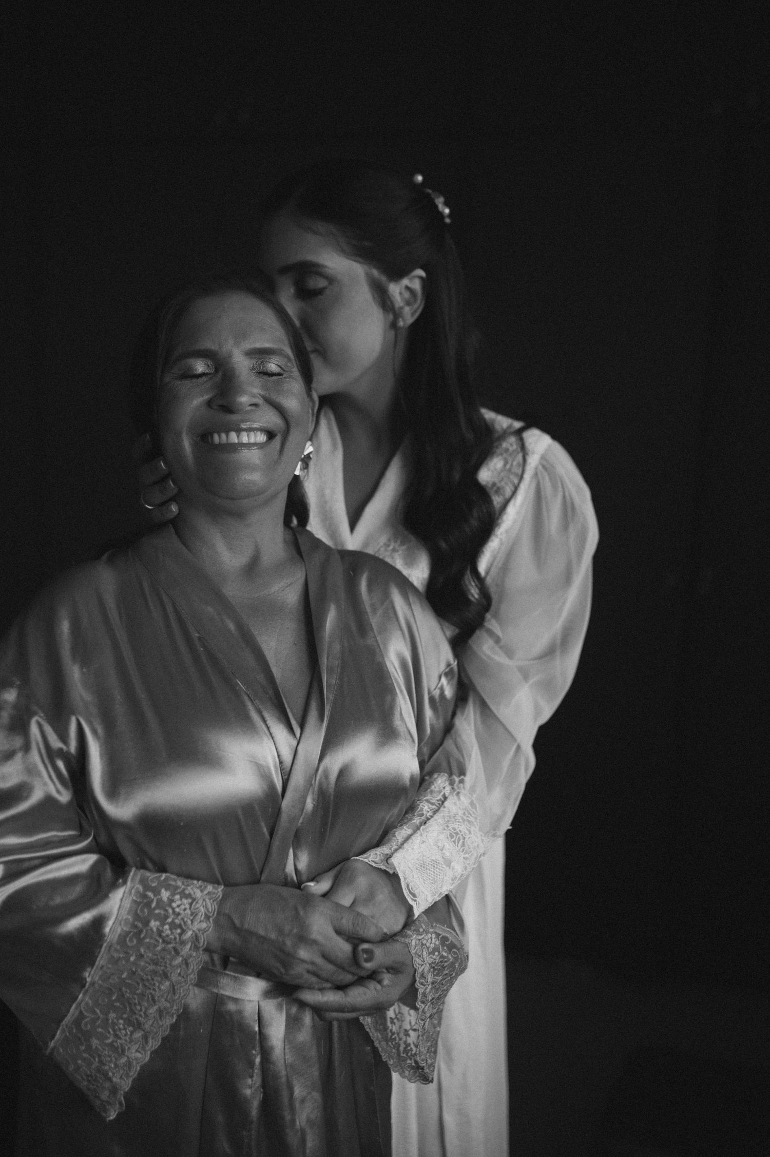 Black and white photo of a bride kissing her smiling mother on the cheek, both wearing satin robes.