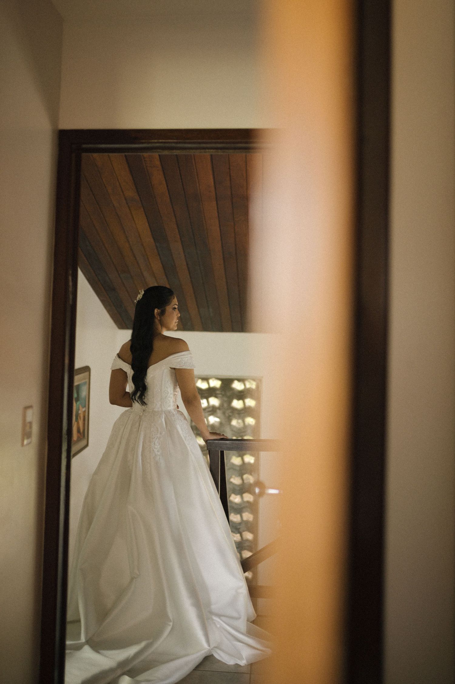 Bride in elegant off-shoulder white ball gown viewed through doorway, standing near vanity mirror with Hollywood lights.