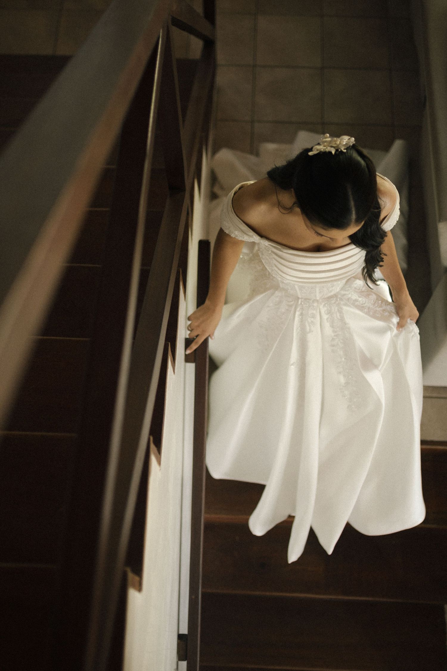 Aerial view of a bride in a white off-shoulder gown descending a wooden staircase, holding her dress.