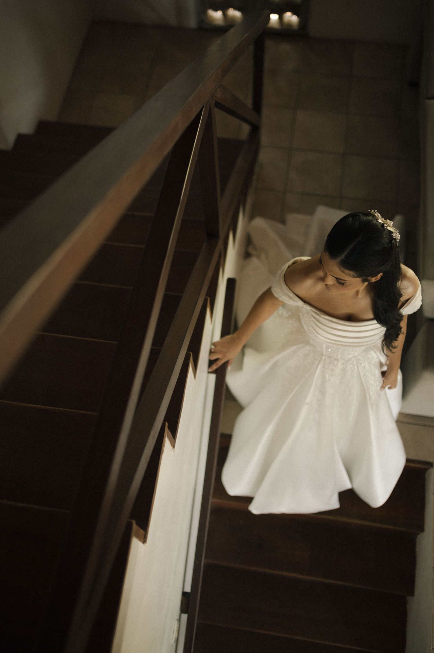 Bride in white wedding gown descending wooden staircase, viewed from above, holding railing elegantly.