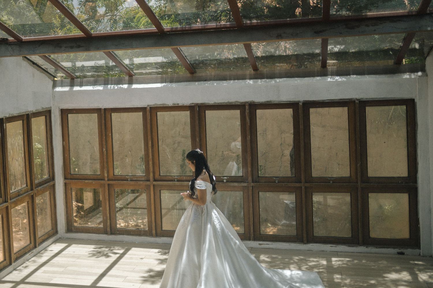 A bride in a white ball gown wedding dress stands in a sunlit glass conservatory with wooden framed windows.