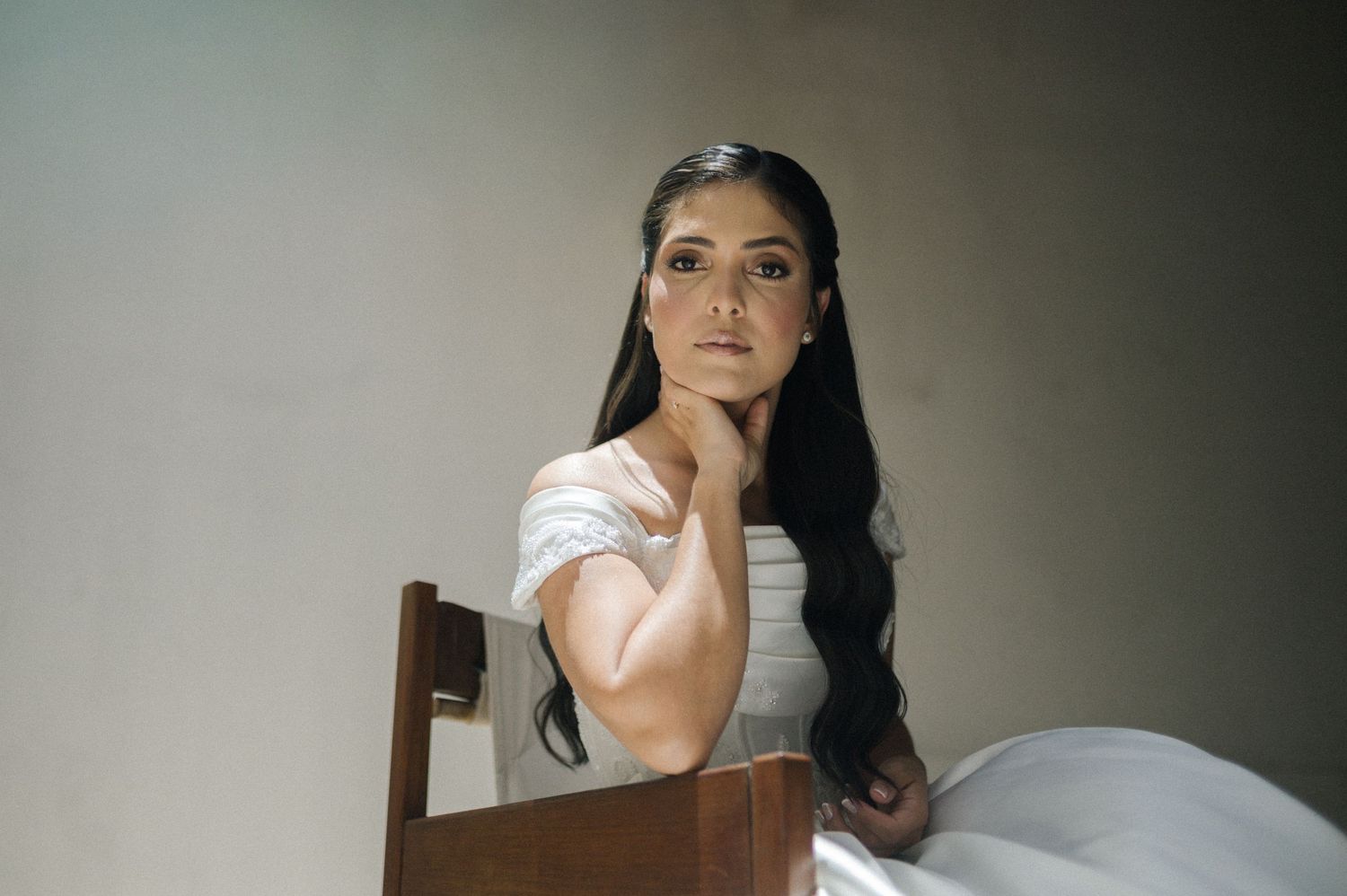Elegant woman in white dress seated on wooden chair, posing thoughtfully with hand on chin against neutral background.