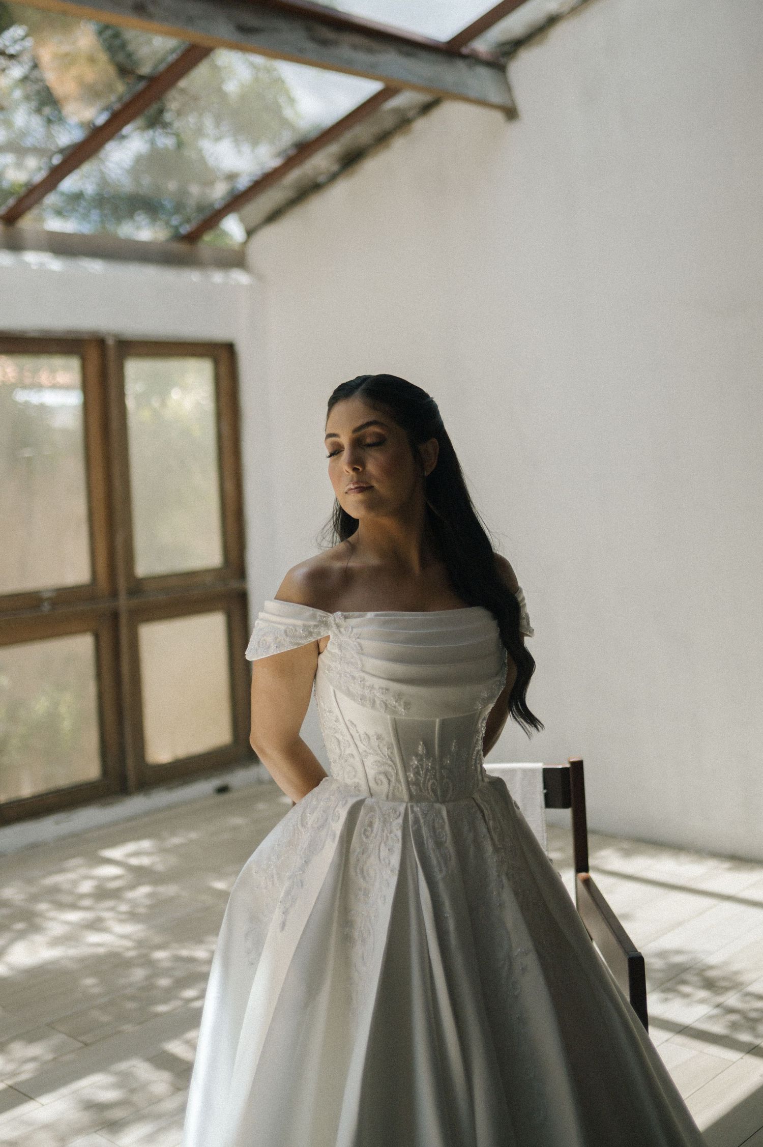 Bride in elegant off-shoulder white ball gown posing in sunlit room with wooden doors.