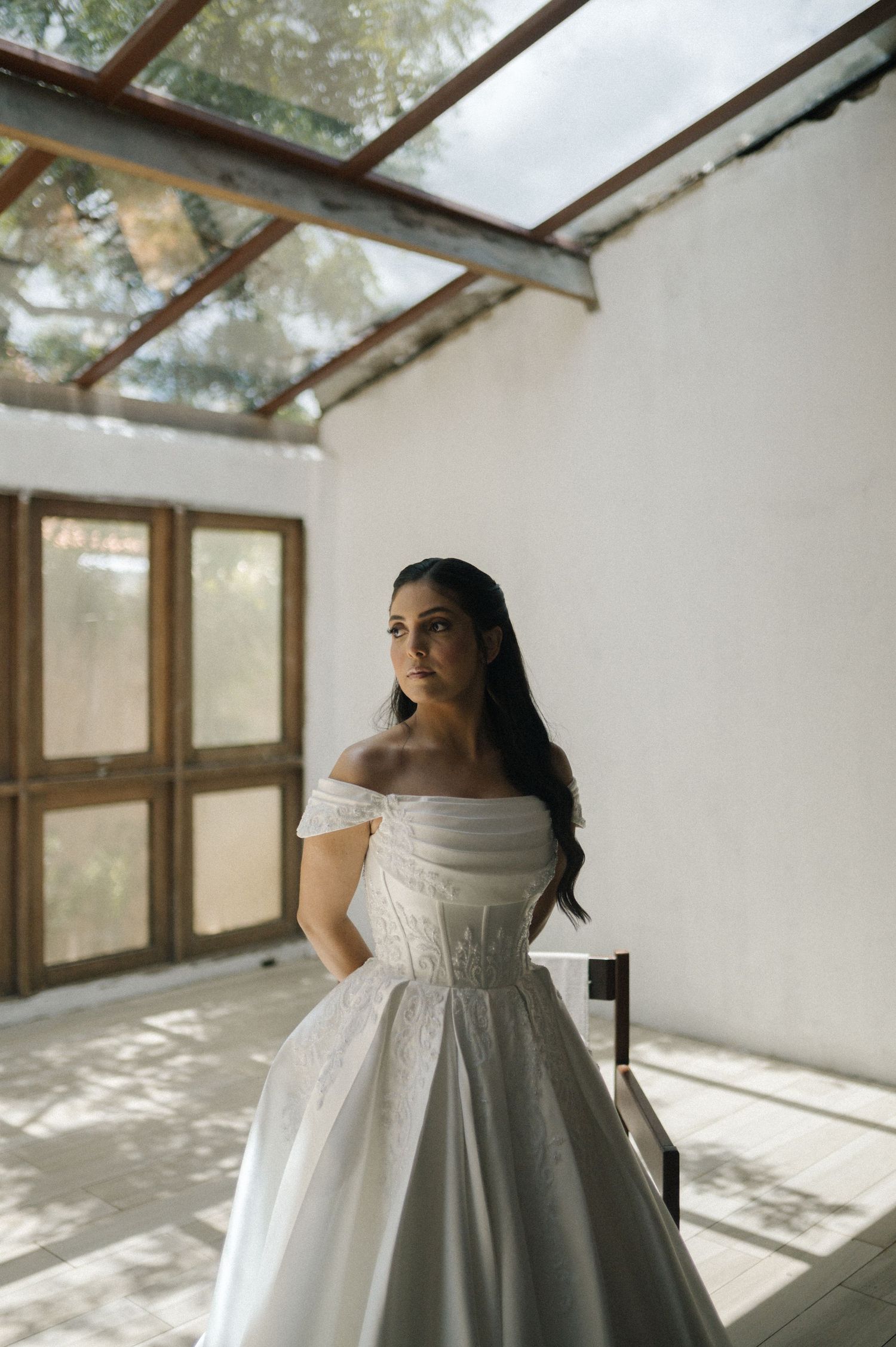 A woman in an off-shoulder white ball gown poses elegantly in a sunlit white room with wooden doors.