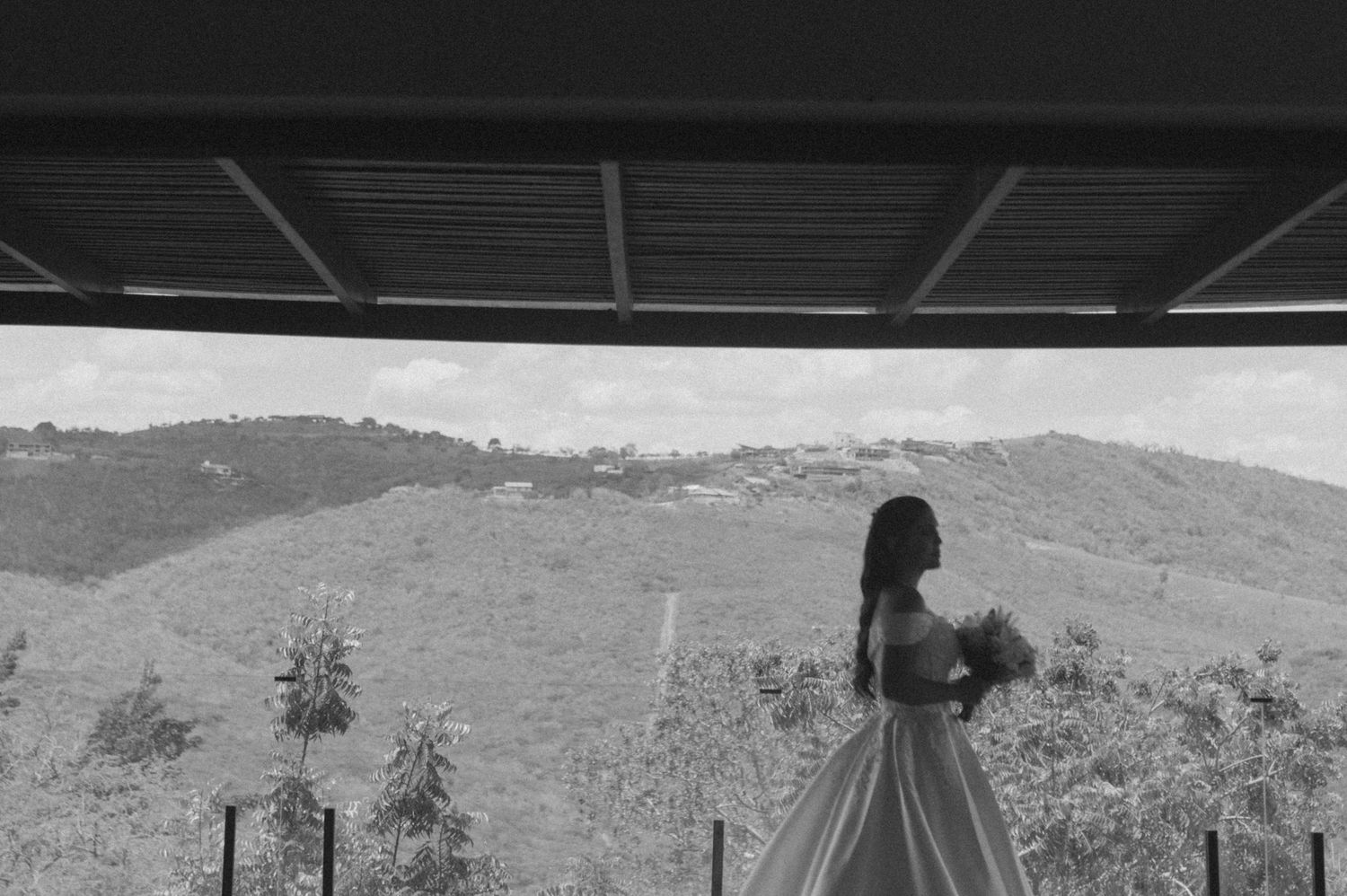 Black and white photo of a bride in a gown holding flowers, gazing at a lush hillside landscape.