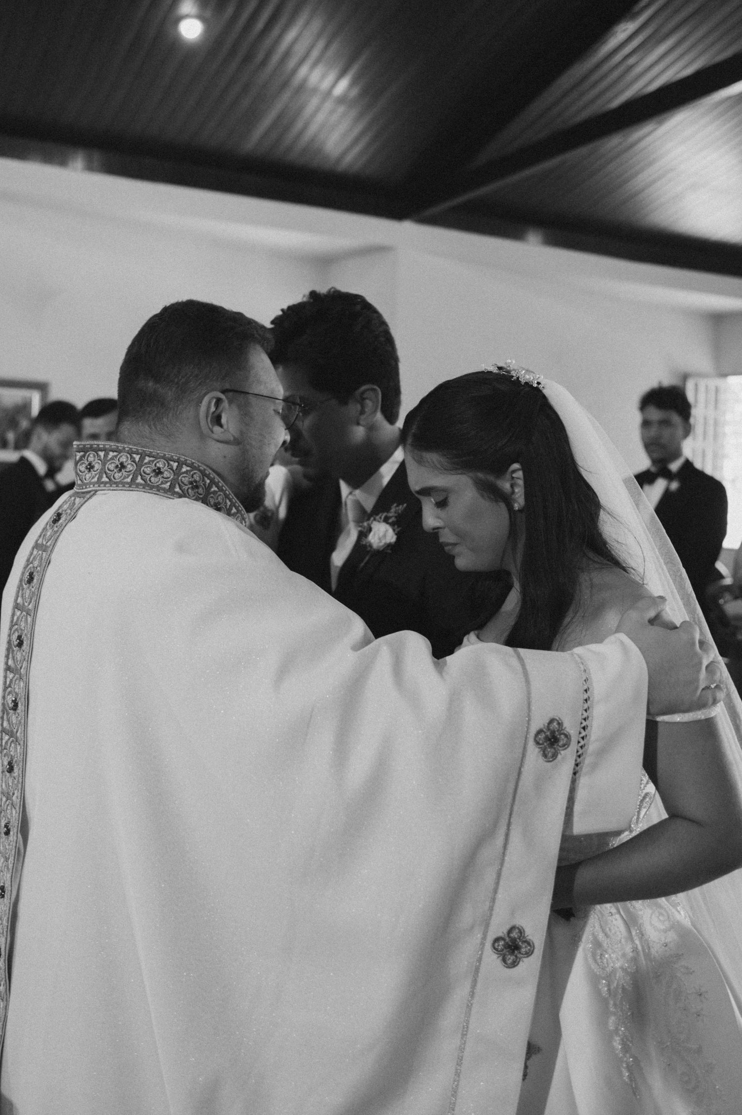 Black and white photo of a bride and groom receiving a blessing from a priest during a church wedding ceremony.