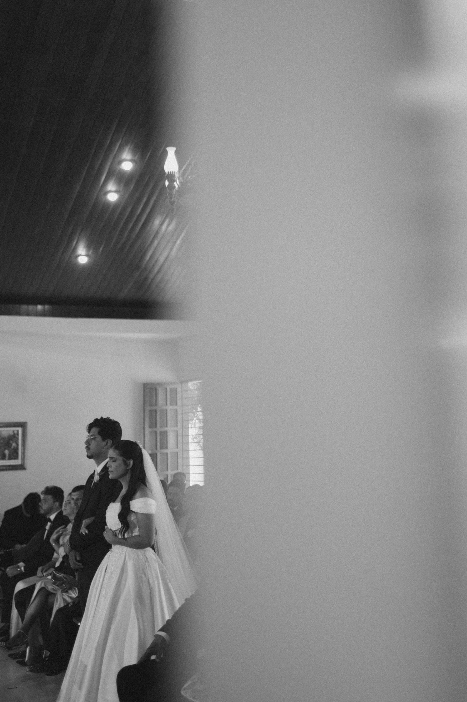 Black and white wedding ceremony photo of bride in white gown and groom standing together inside a chapel.