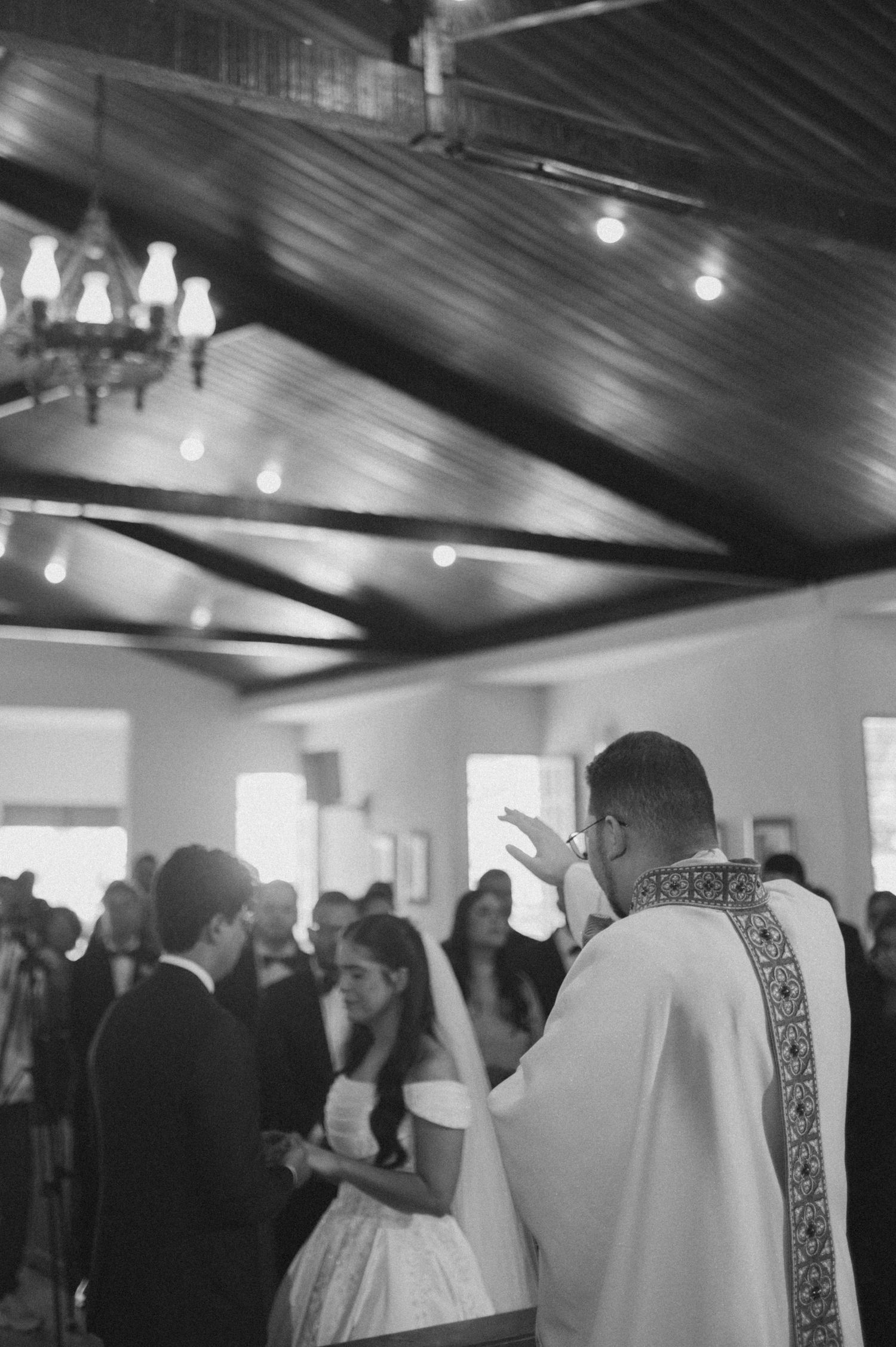 Black and white photo of a priest officiating a wedding ceremony in a rustic church with wooden ceiling beams.