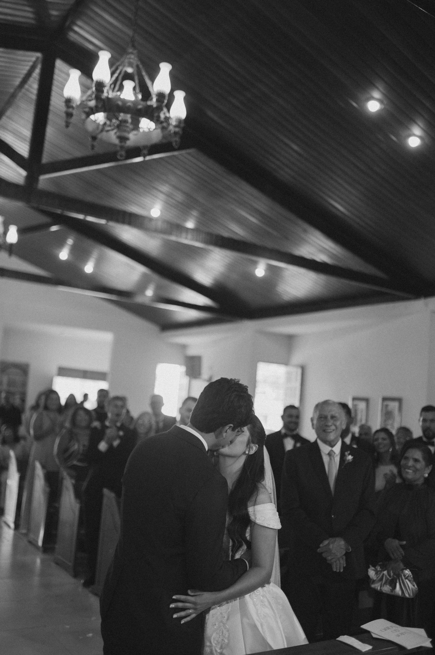 Black and white photo of a couple sharing their first kiss at a wedding ceremony in a rustic chapel with guests watching.