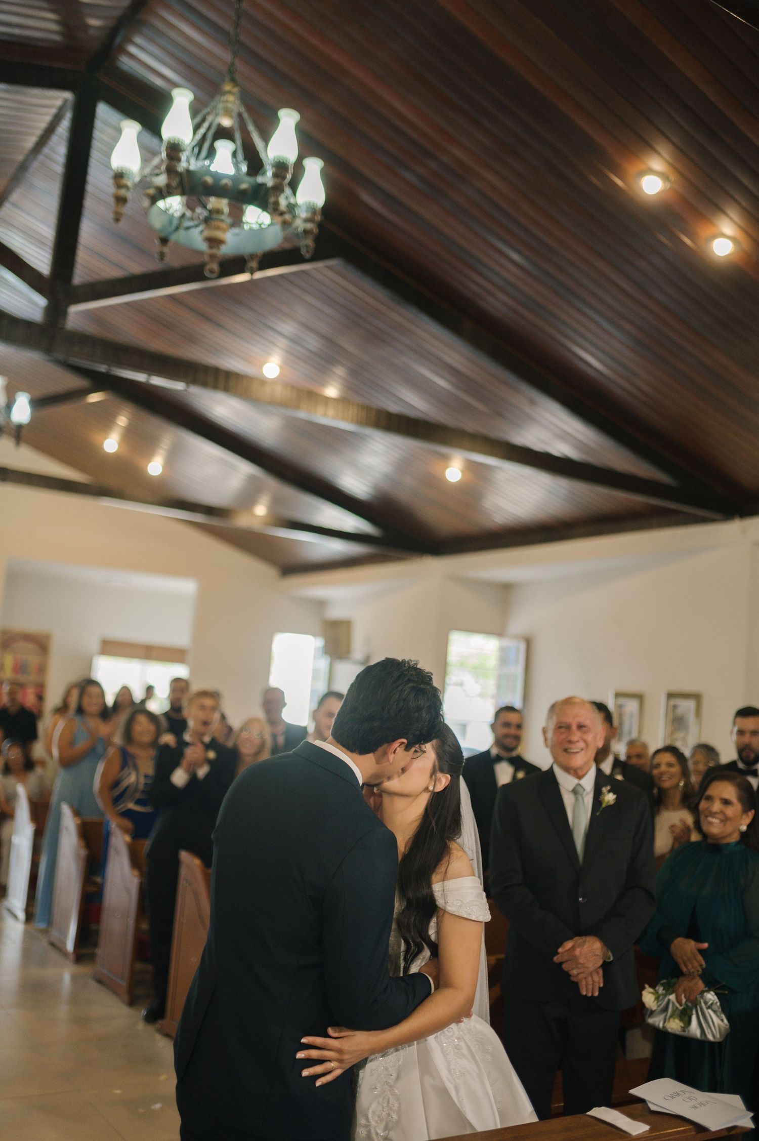 Bride and groom share first kiss at wedding ceremony altar with guests applauding in elegant chapel venue.