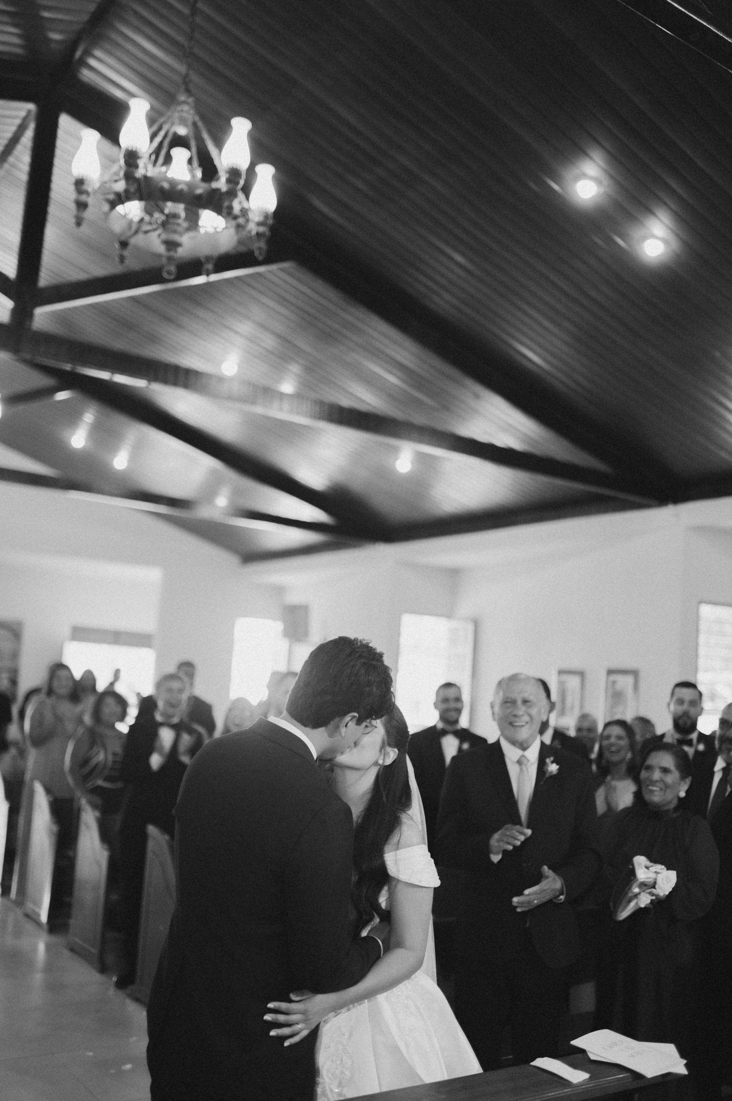 Black and white photo of a couple sharing their first kiss at a wedding ceremony in a chapel.