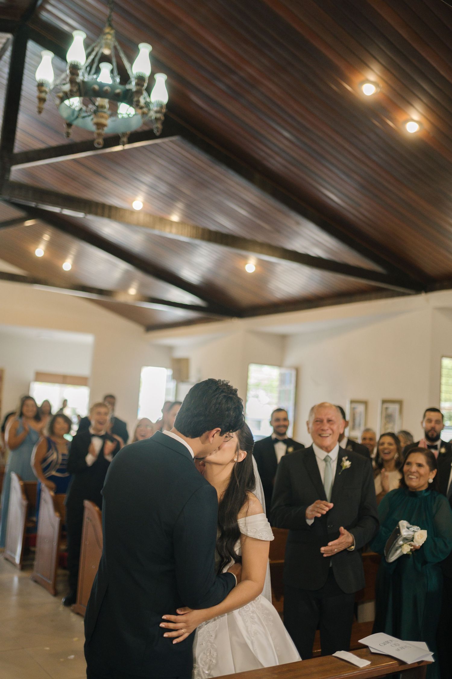 Bride and groom share first kiss at church altar while smiling guests watch during elegant wedding ceremony.