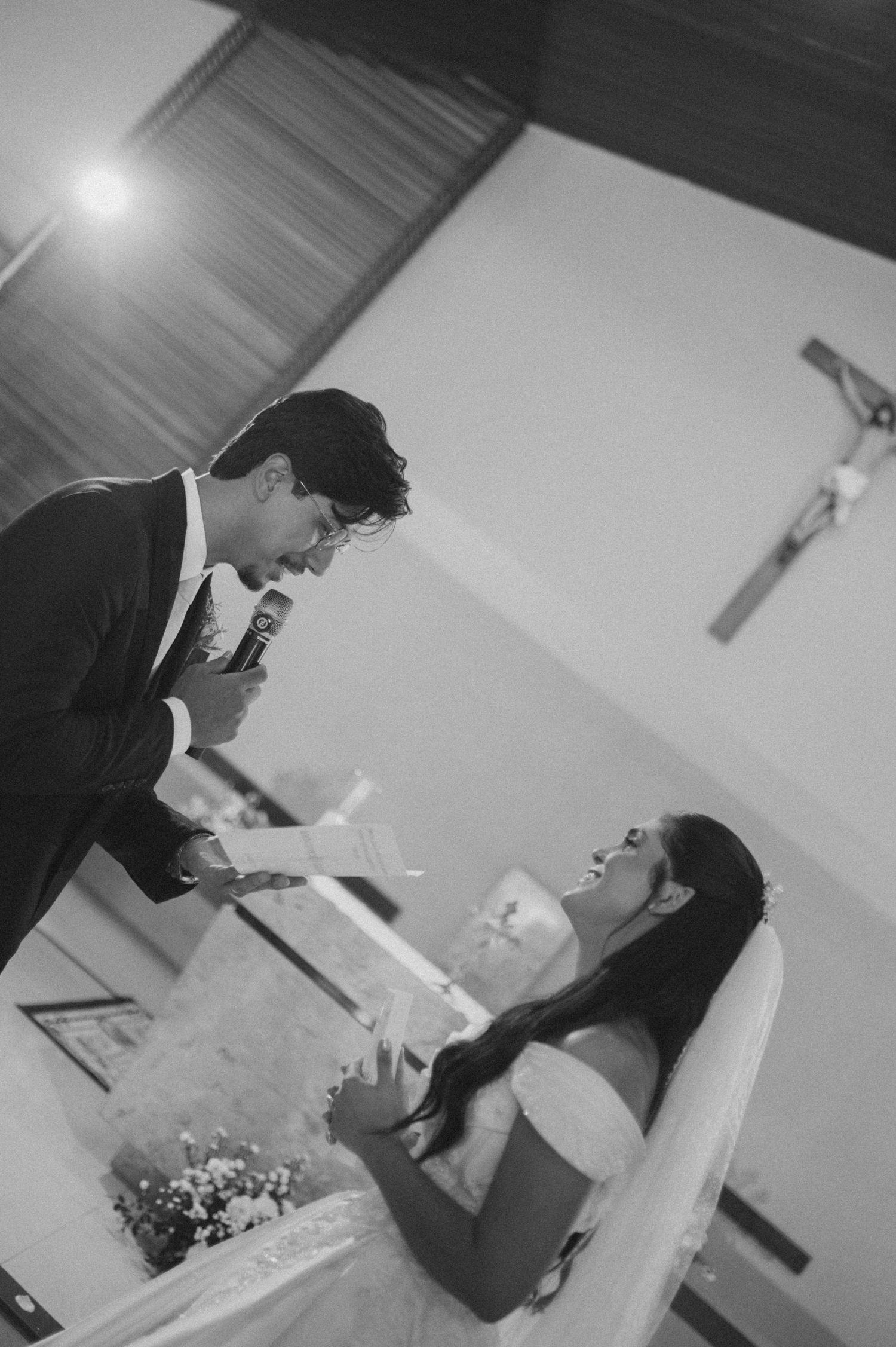 Black and white wedding ceremony photo of groom reading vows to bride at church altar with crucifix in background.
