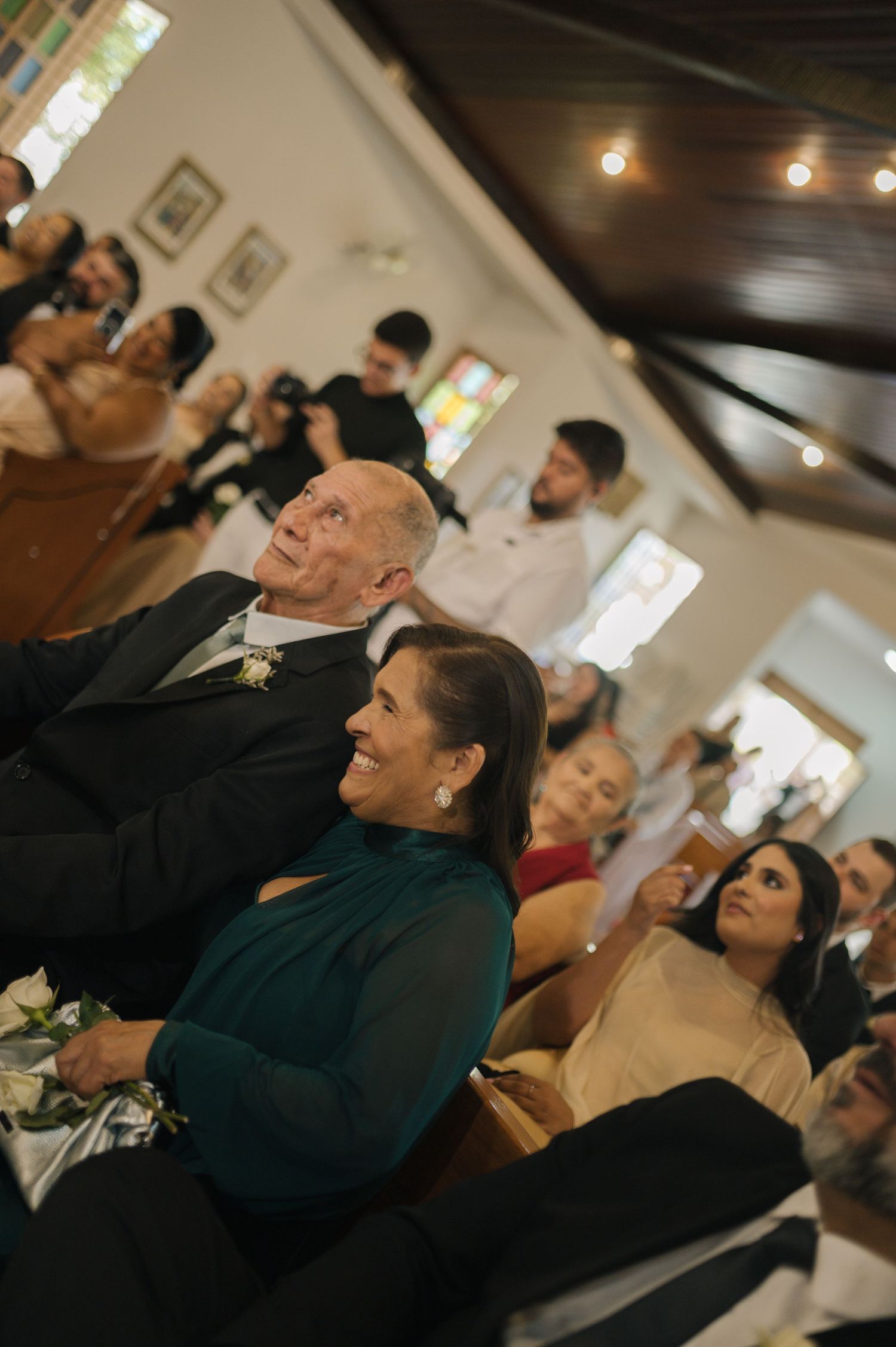 Wedding guests smile and watch ceremony in a warmly lit chapel, woman in teal dress seated beside older man in suit.