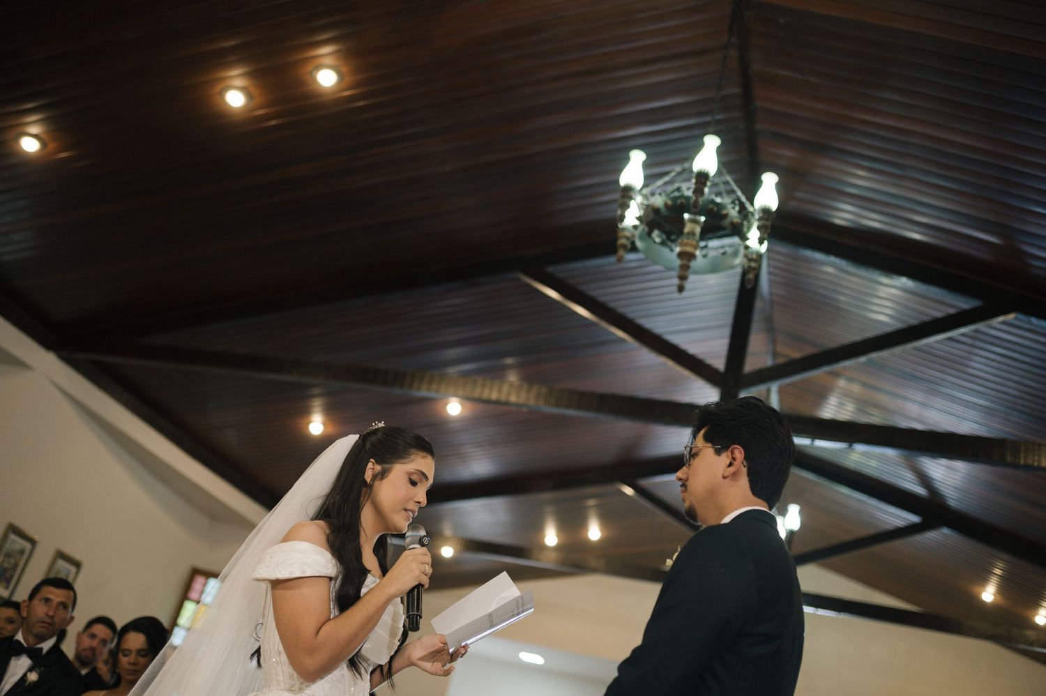 Bride reads wedding vows at indoor ceremony with chandelier lighting and groom standing opposite in formal attire.