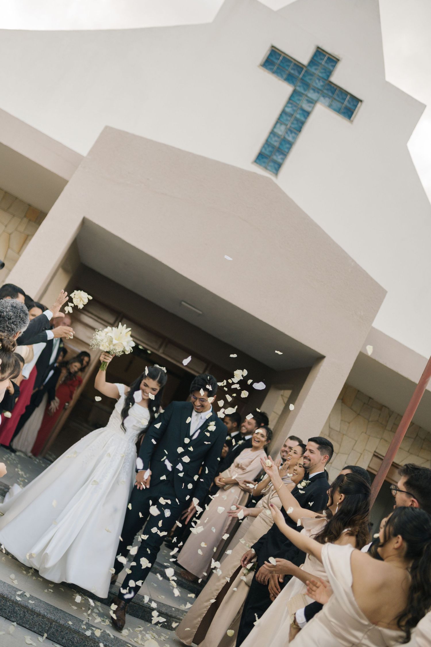 Bride and groom exit white chapel with cross, surrounded by wedding party tossing flower petals in celebration.