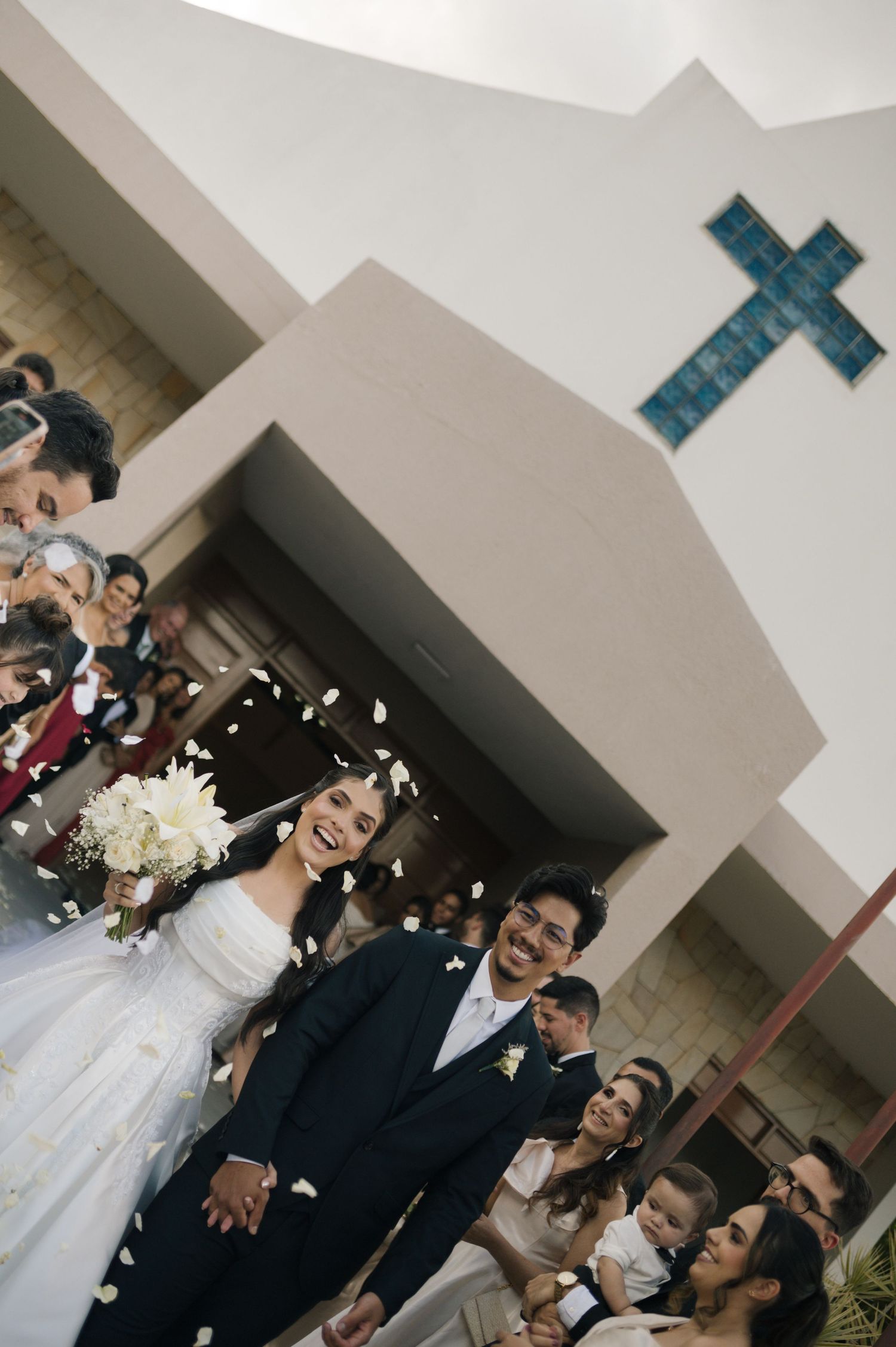 Joyful newlyweds exit a white church as guests toss flower petals in celebration.