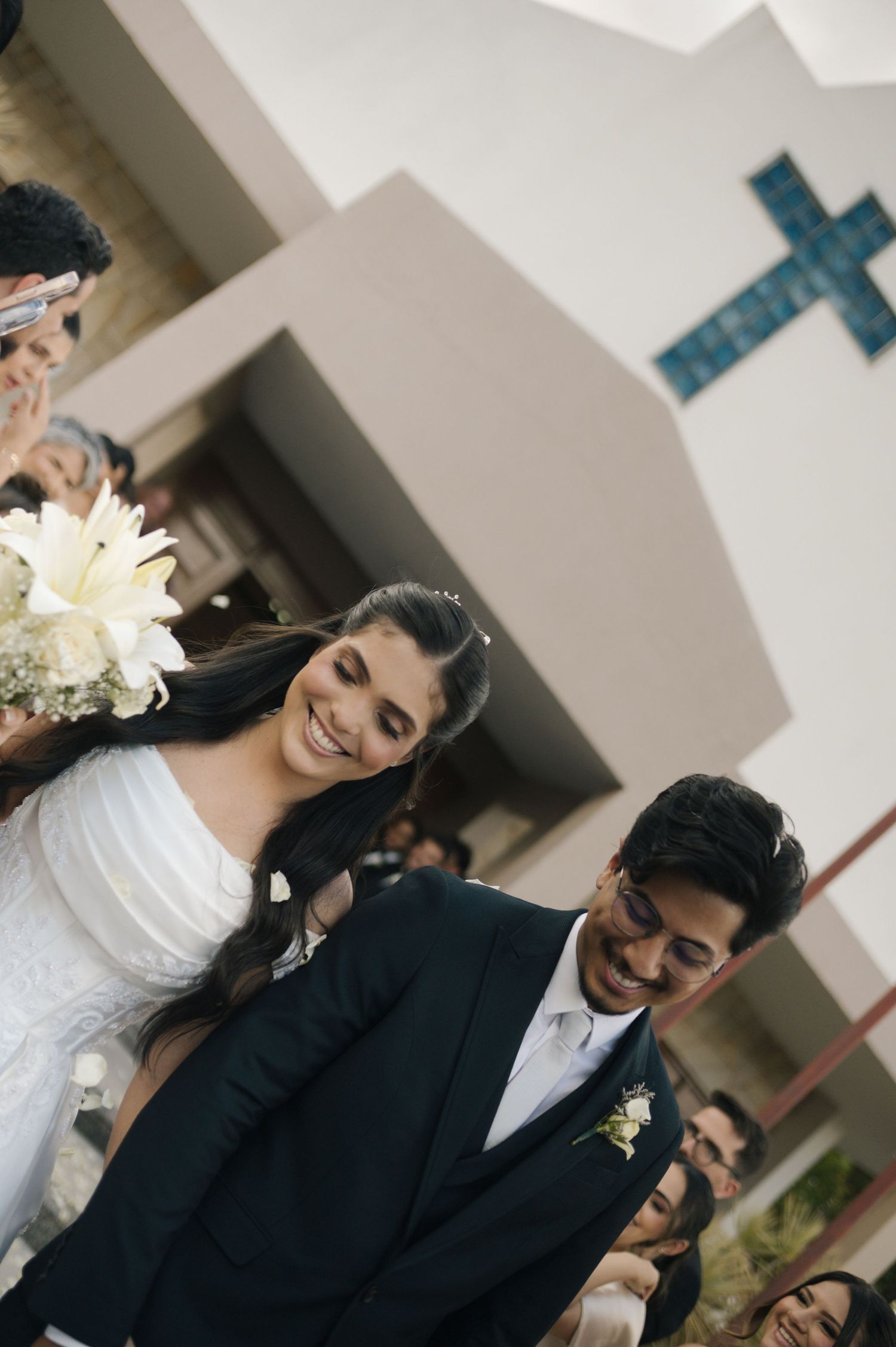 Smiling bride in white gown holding bouquet walks with groom in black suit outside church with blue cross.