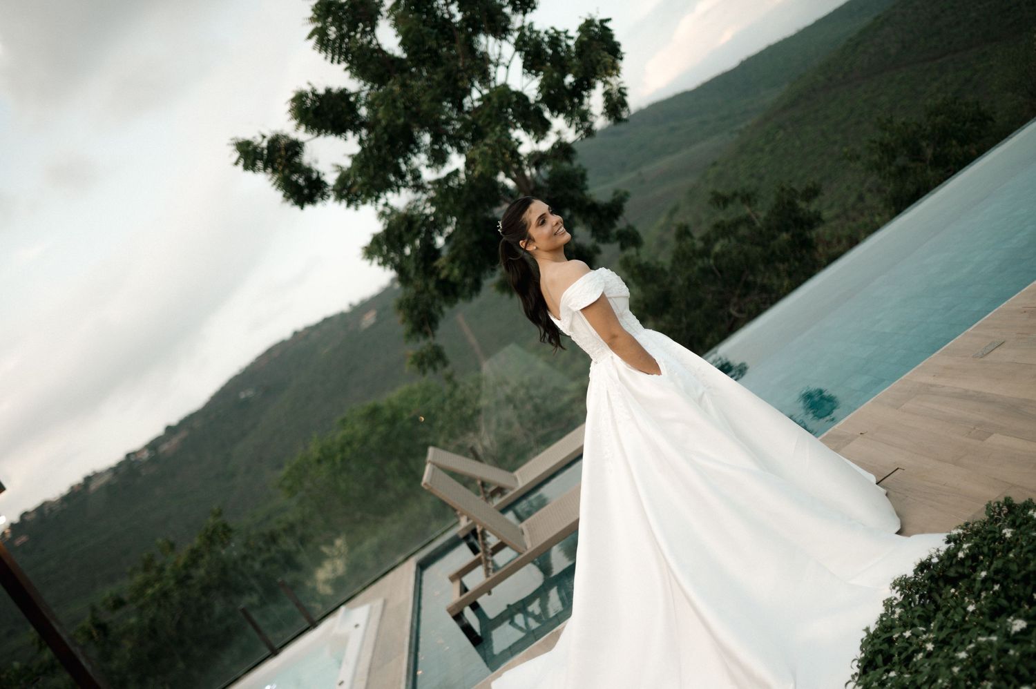 Elegant bride in flowing white ball gown poses on balcony overlooking lush green mountain landscape.