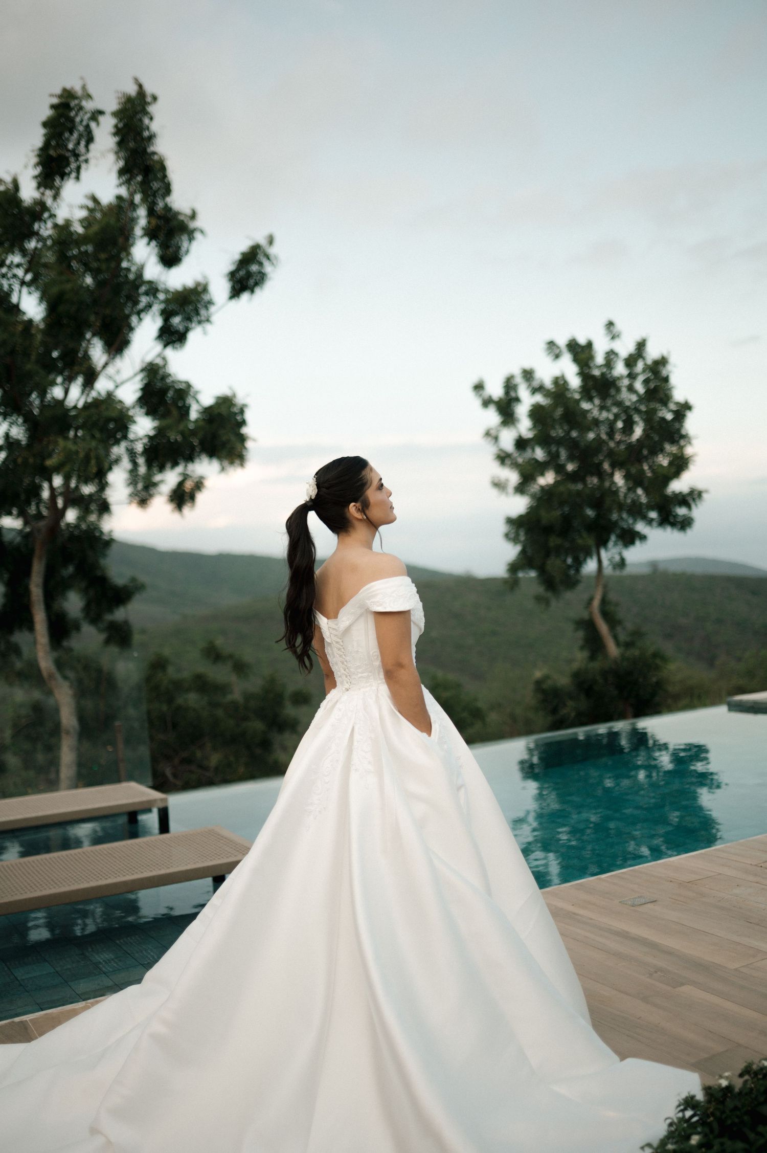 Bride in elegant off-shoulder white ball gown stands by infinity pool with mountain views at dusk.