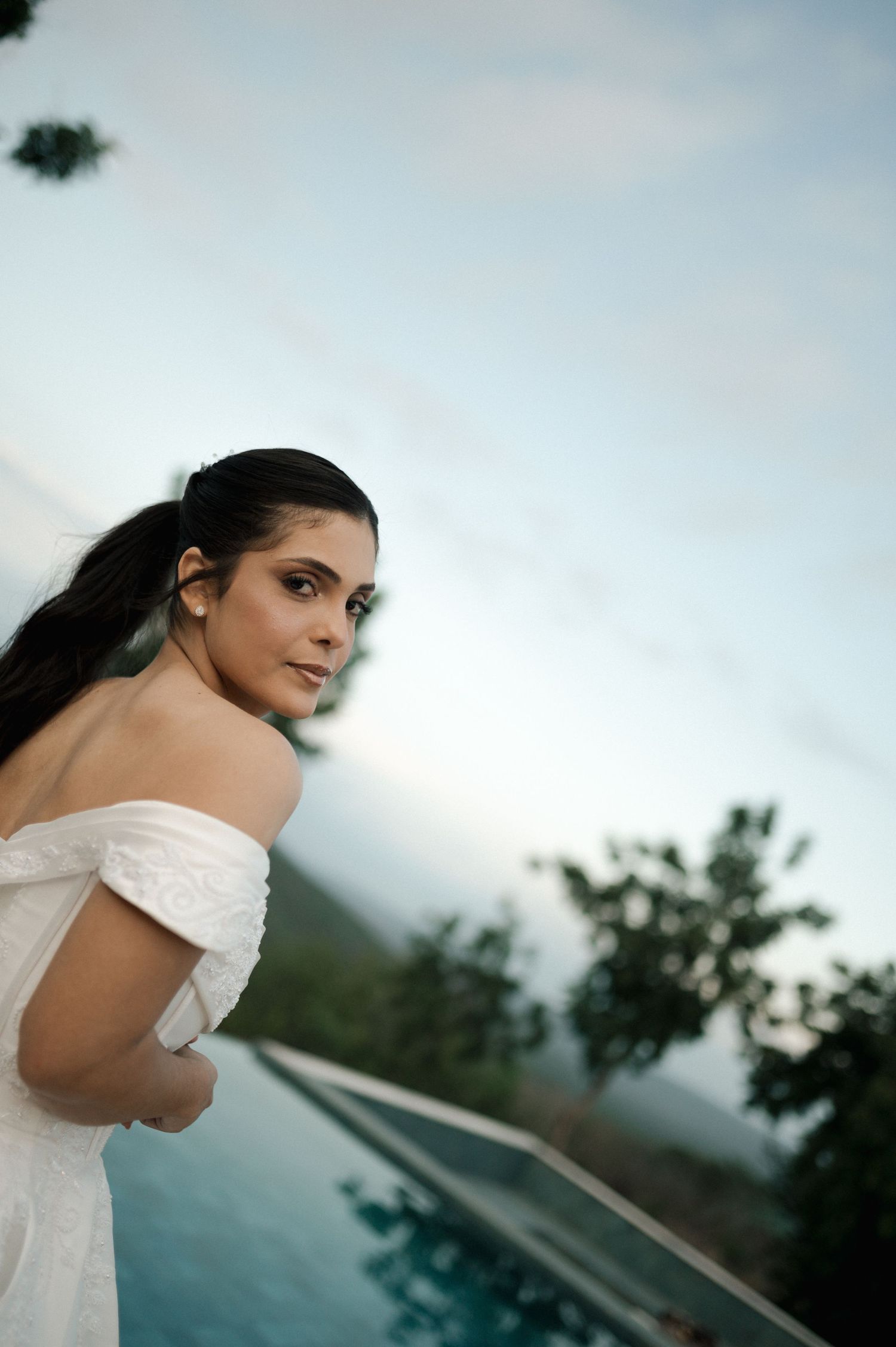Beautiful bride in off-shoulder white dress gazes back over shoulder beside infinity pool at dusk.